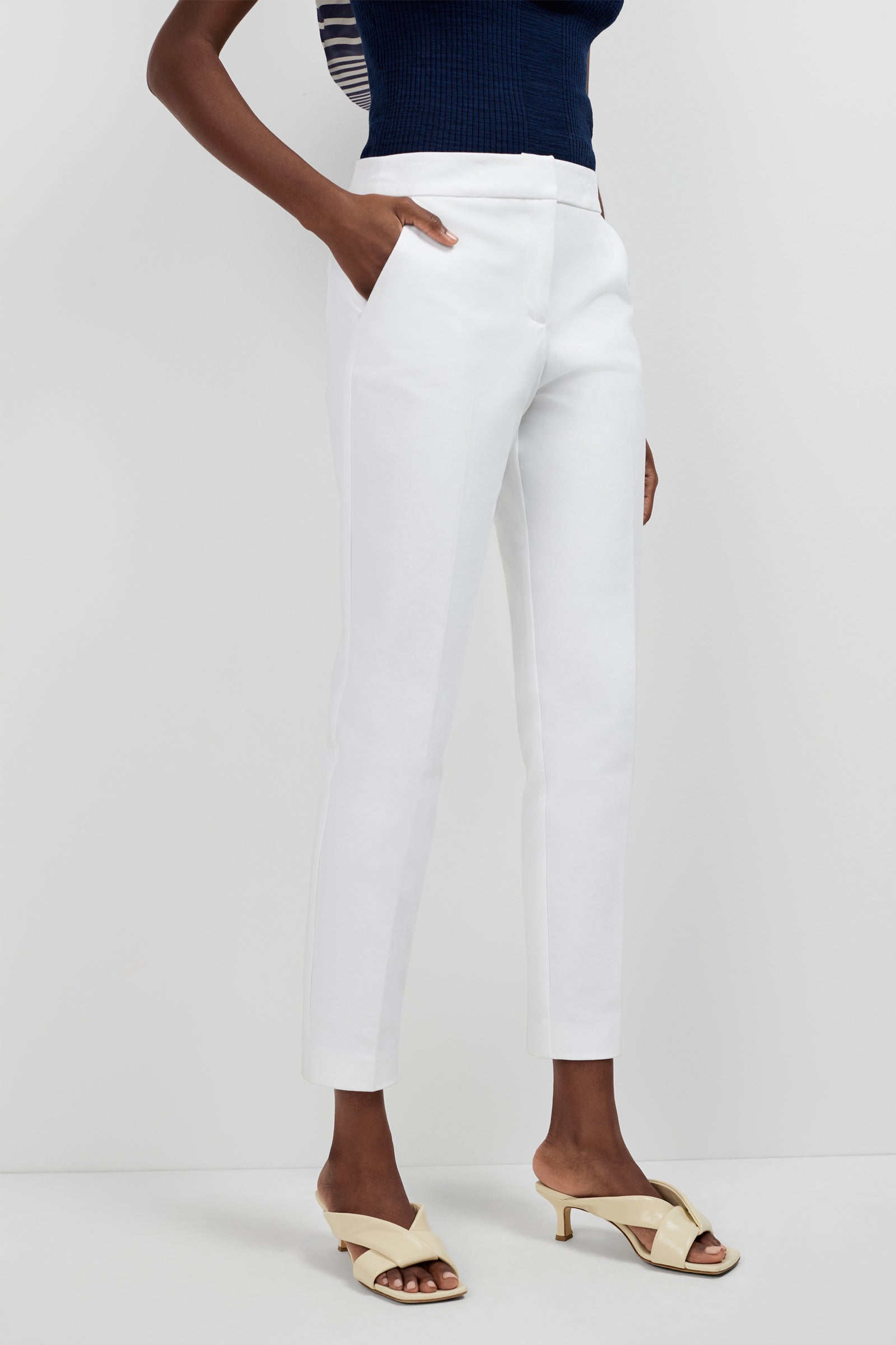 A person’s lower body is shown against a white background, wearing white Aubree 2 Cigarette Pants, a navy sleeveless top, and cream heeled sandals.