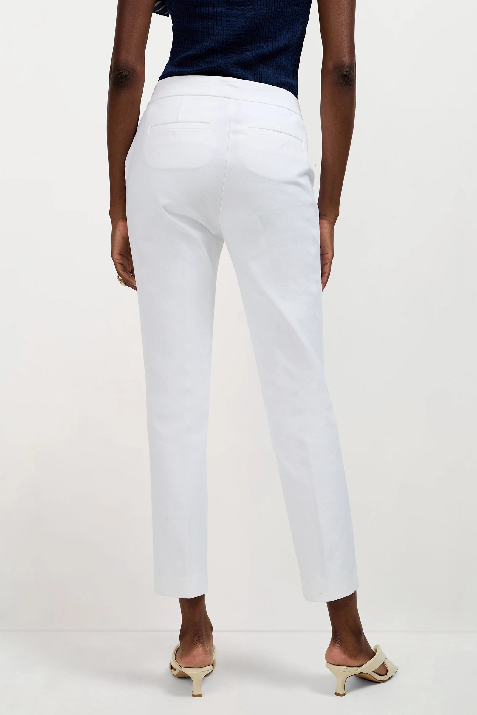 A person is shown from the back wearing white Aubree 2 Cigarette Pants, a dark sleeveless top, and cream heeled sandals against a plain light background.