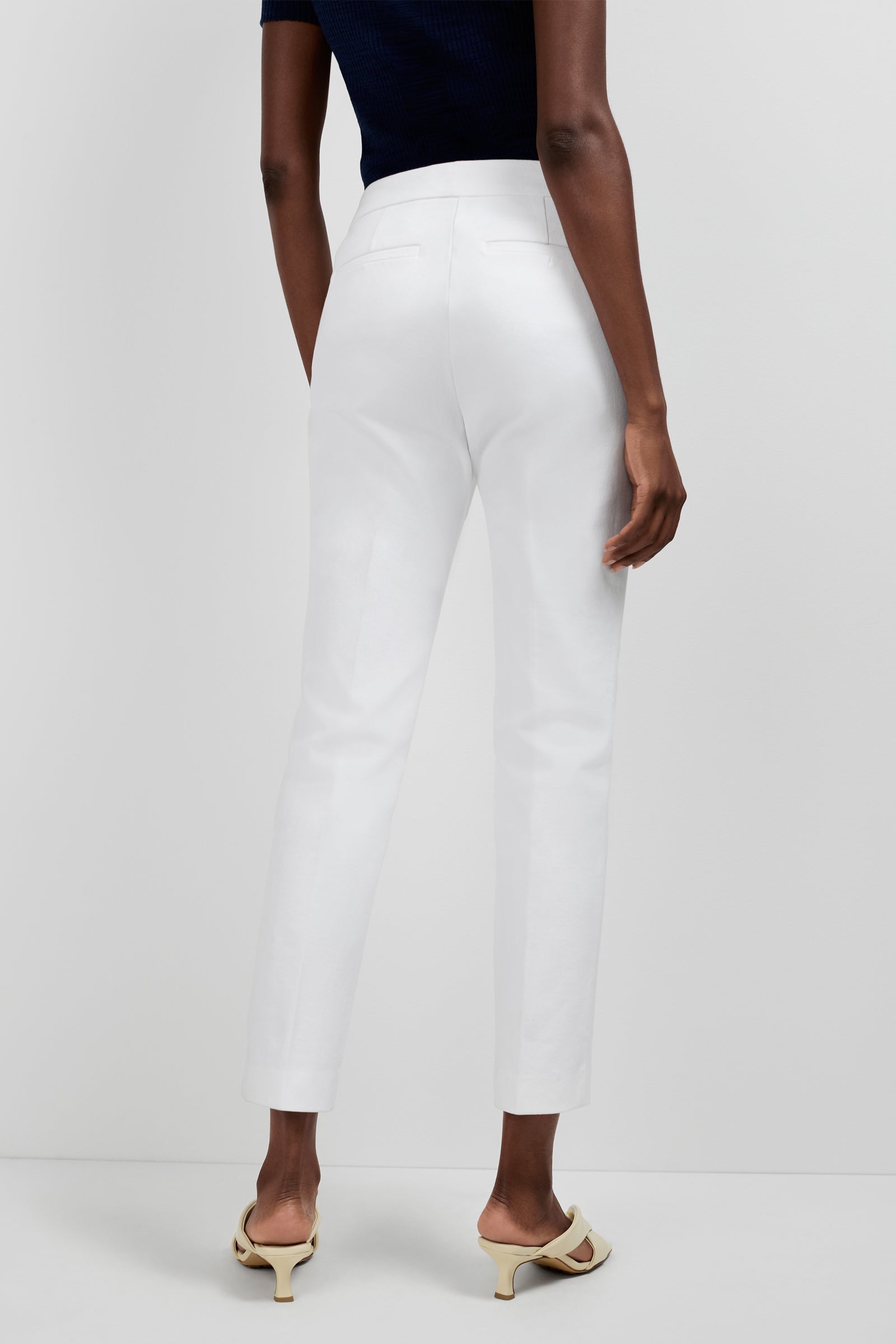 A person stands against a plain background, shown from the back, wearing a short-sleeve black top, white Aubree 2 Cigarette Pants, and light-colored open-toe heeled sandals.