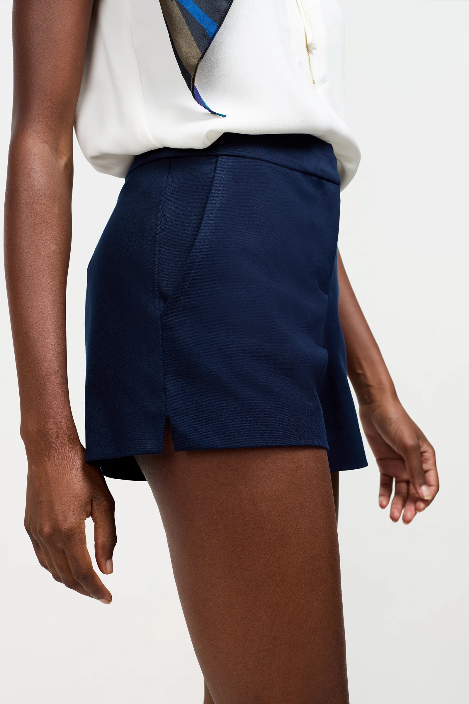 A person wearing a sleeveless white top with patterned detail and the Corbin Shorts - Cotton Bi-Stretch in navy blue, standing against a plain light background.