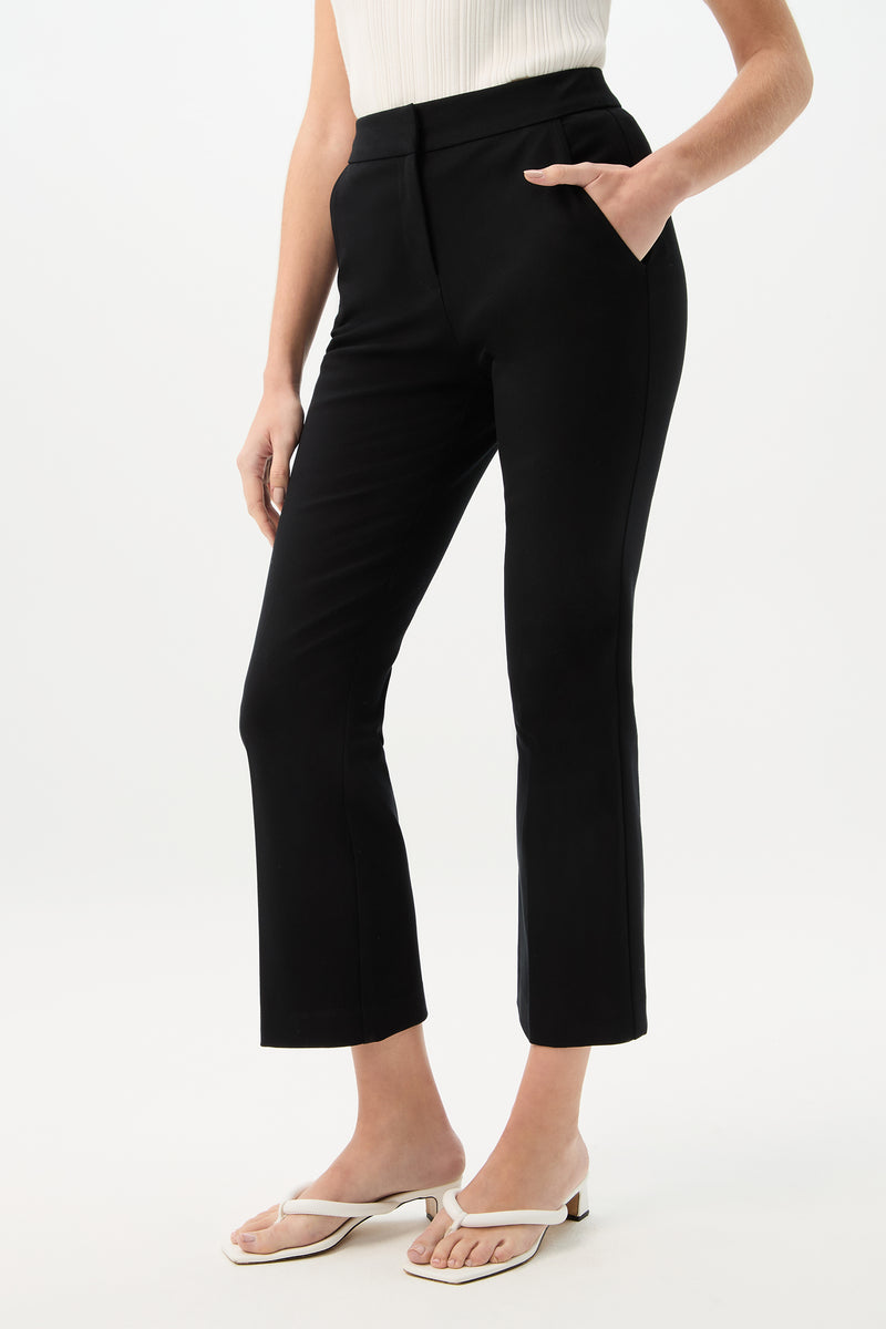 A person wearing the Lulu Cropped Pant - Cotton Bi-Stretch in black, paired with a sleeveless white top and white sandals, stands with one hand in their pocket against a plain white background.