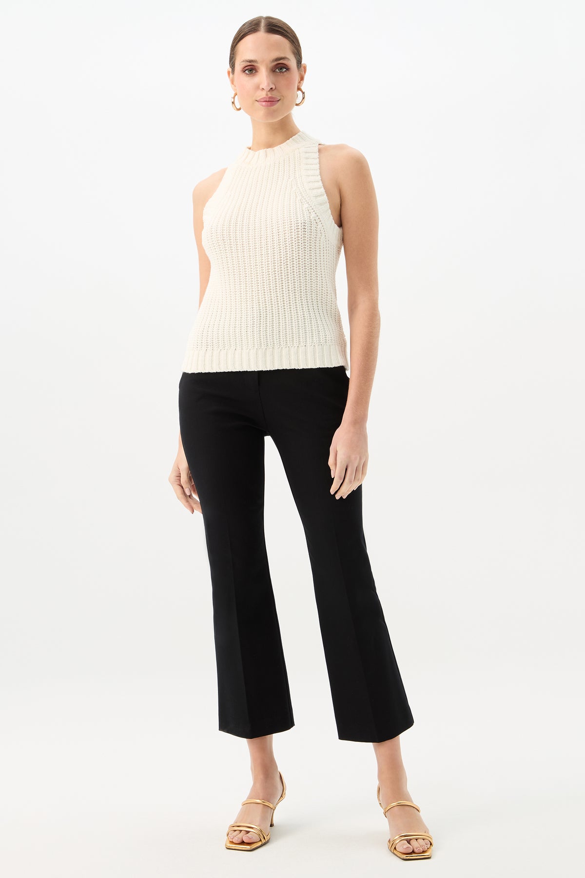 A woman stands against a white background wearing a sleeveless, ribbed cream knit top, beige heeled sandals, hoop earrings, and the Lulu Cropped Pant - Cotton Bi-Stretch in black; her hair is pulled back.
