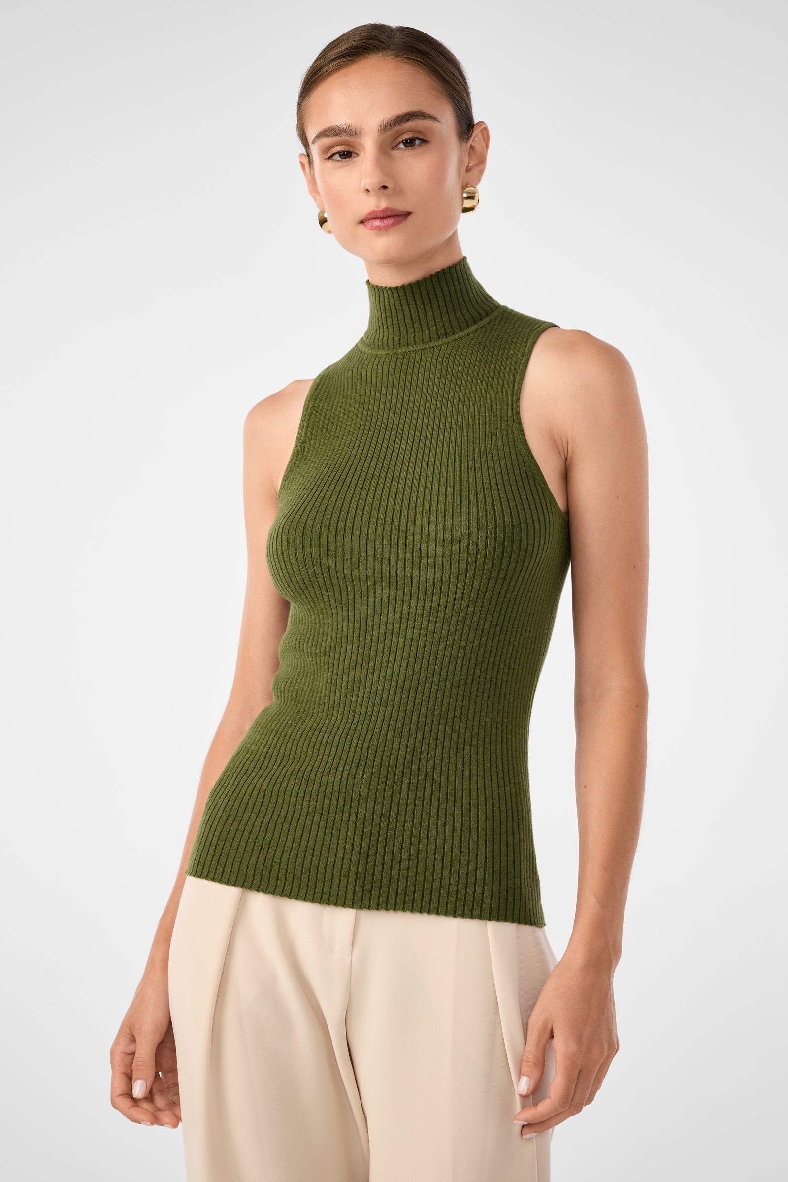 A woman in the Westhampton Knit Pullover in Wilderness pairs it with high-waisted light beige pants. She stands against a light gray background, sporting short, slicked-back hair and gold hoop earrings.