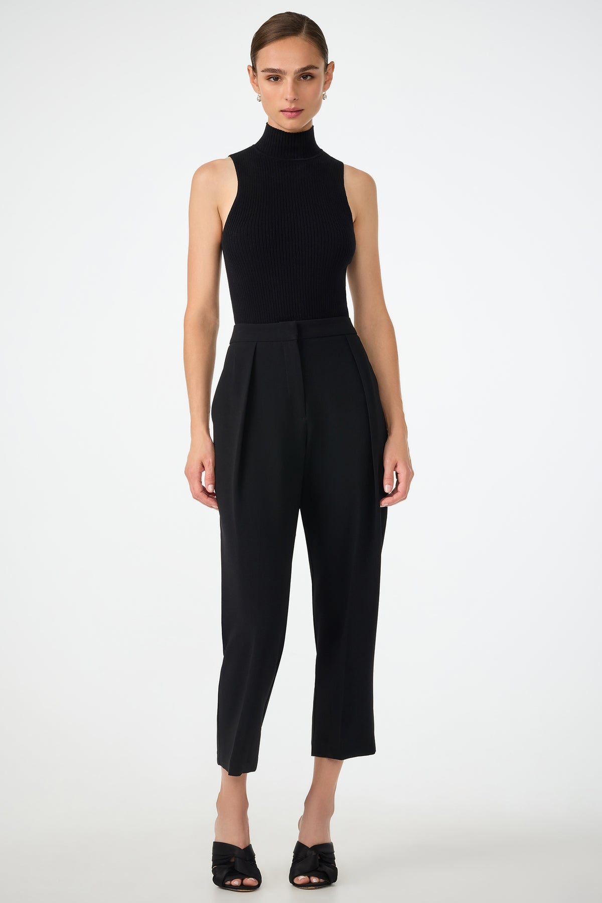 A woman stands against a plain white background, wearing the sleeveless black Westhampton Knit Pullover with high-waisted black tailored pants and open-toe heels. Her hair is pulled back and she has a neutral expression.