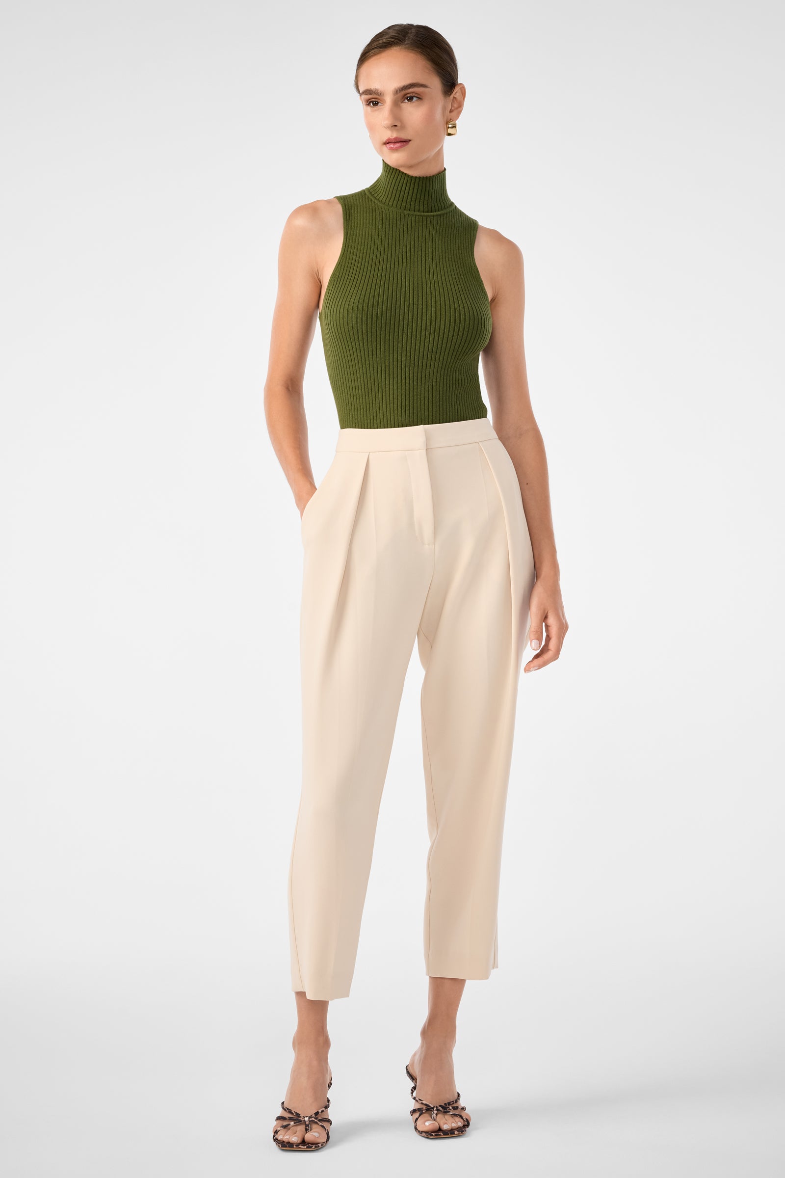 A woman stands against a plain background wearing the Westhampton Knit Pullover in olive green, styled with high-waisted cream pleated pants and strappy heels; her hair is pulled back.