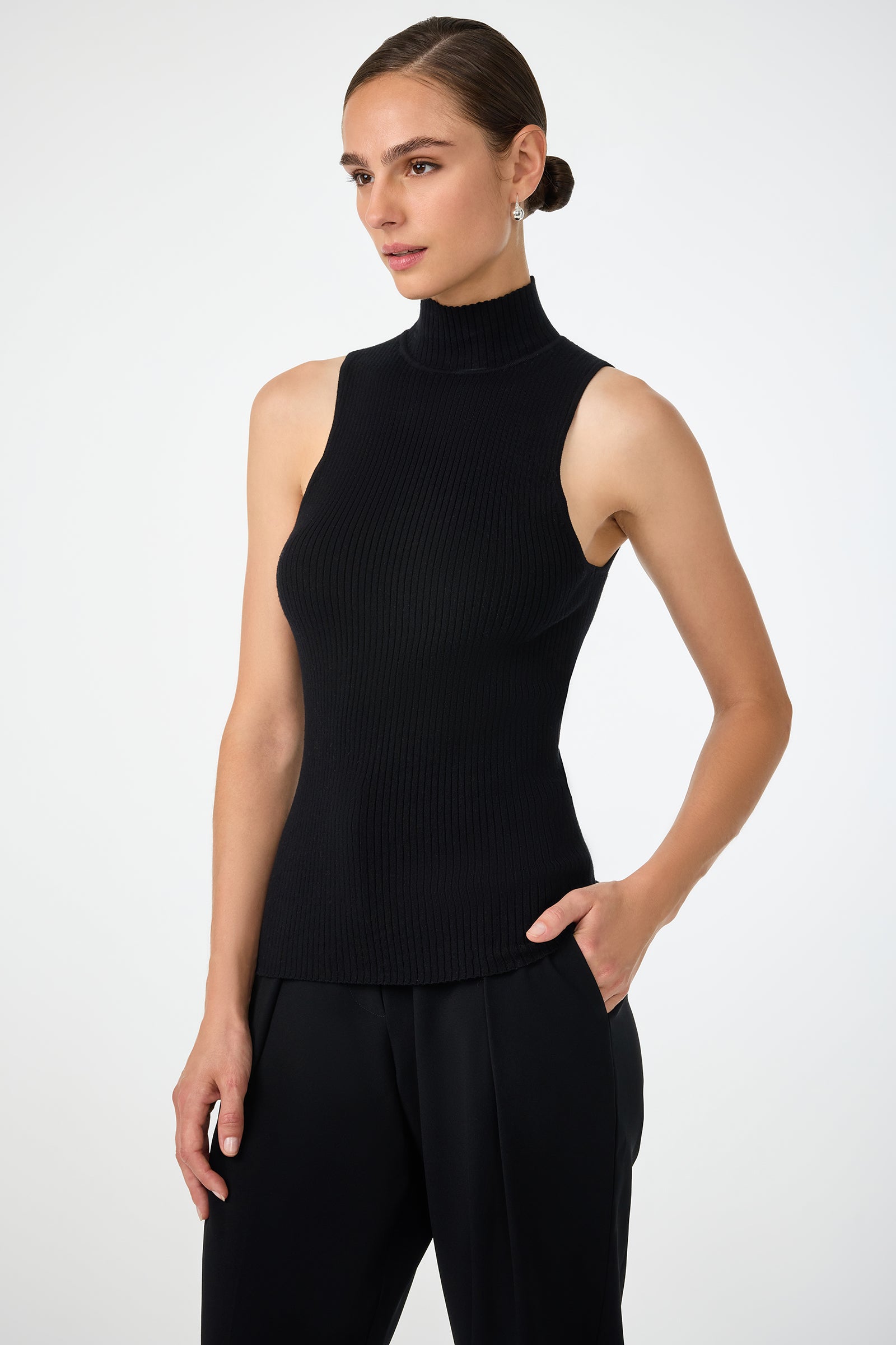 A woman stands against a plain white background wearing the Westhampton Knit Pullover—a sleeveless, black ribbed turtleneck top—paired with black pants and one hand in her pocket.