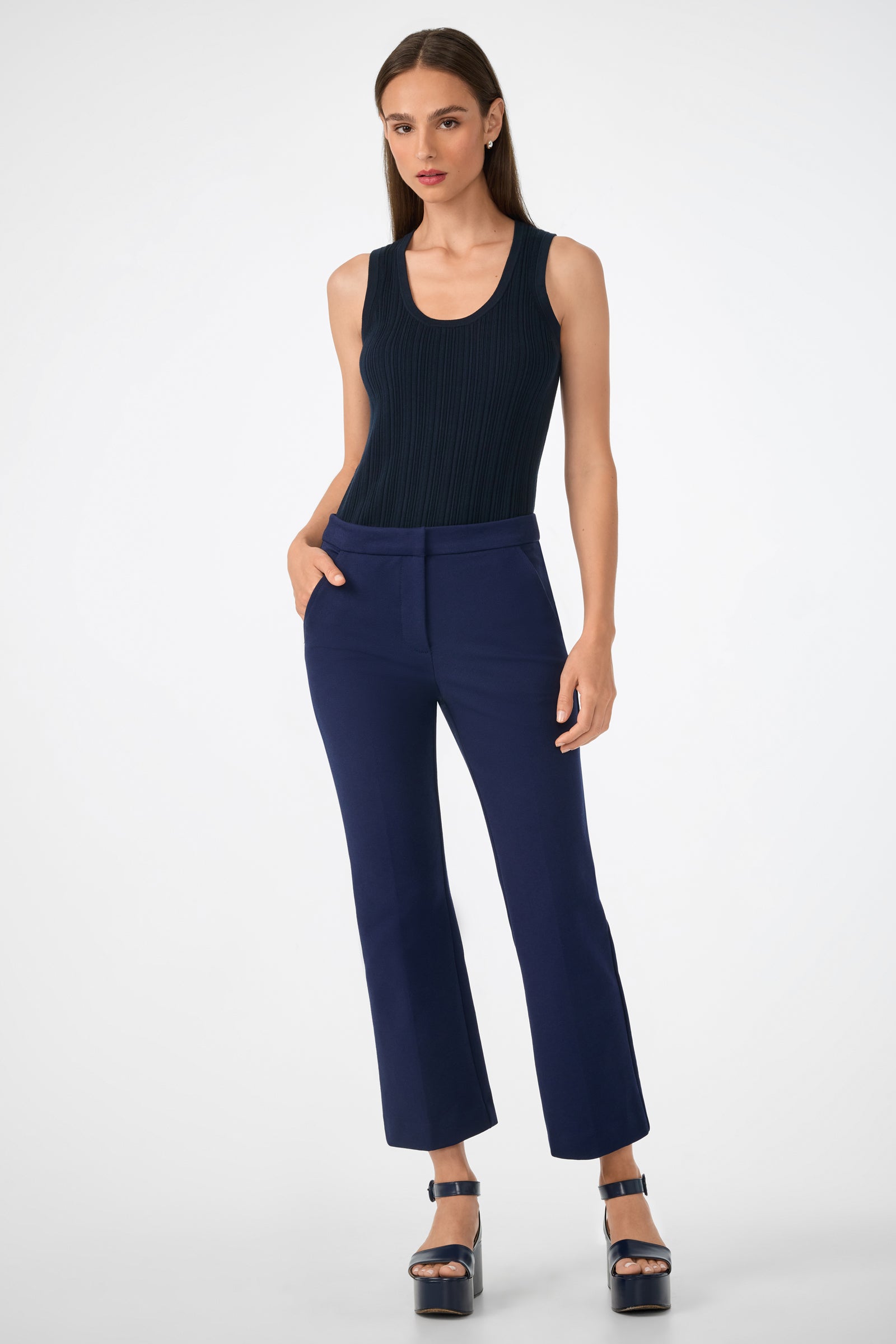 A woman with straight brown hair stands against a white background wearing navy blue Banshee Pant, a sleeveless black ribbed top, and navy blue platform sandals. Her hands are in her pockets.