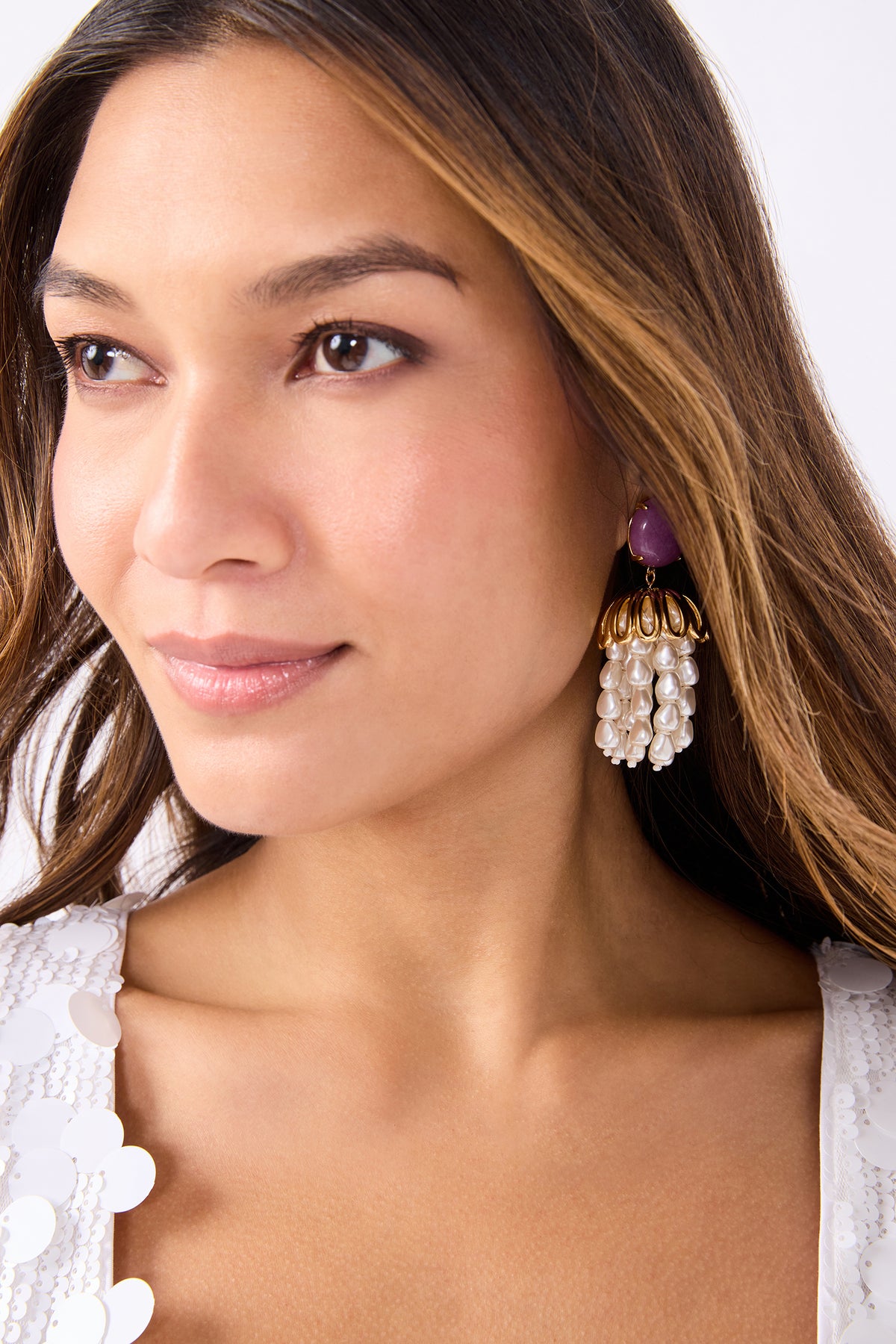 A woman with long brown hair wears a white sequin top and large Lele Sadoughi x Trina Turk Parapluie Strand Earrings with pearls, gold accents, and a purple stone. She gazes slightly to the side with a soft, neutral expression.