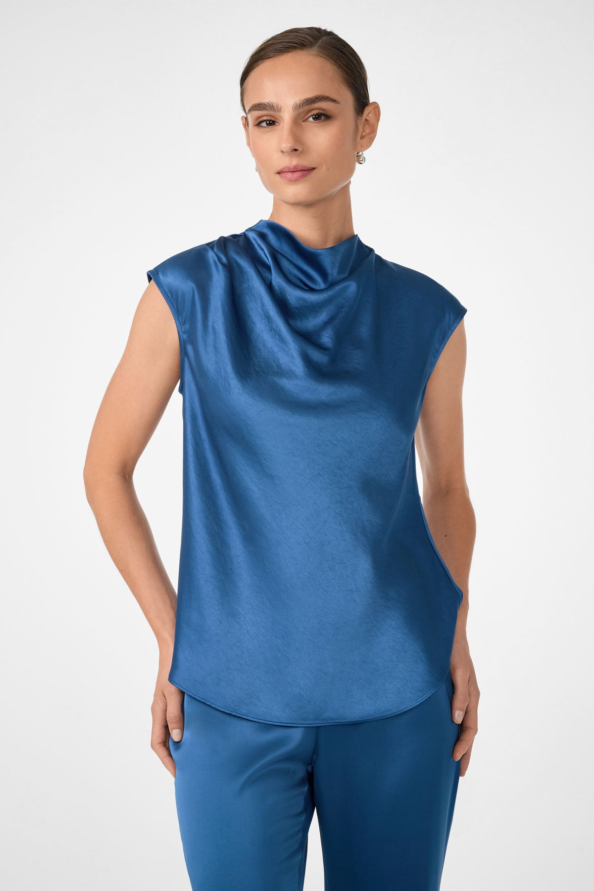 A woman confidently poses against a light background, wearing the Mikan Sleeveless Top in Sumi Blue with a draped neckline, paired with matching blue pants.