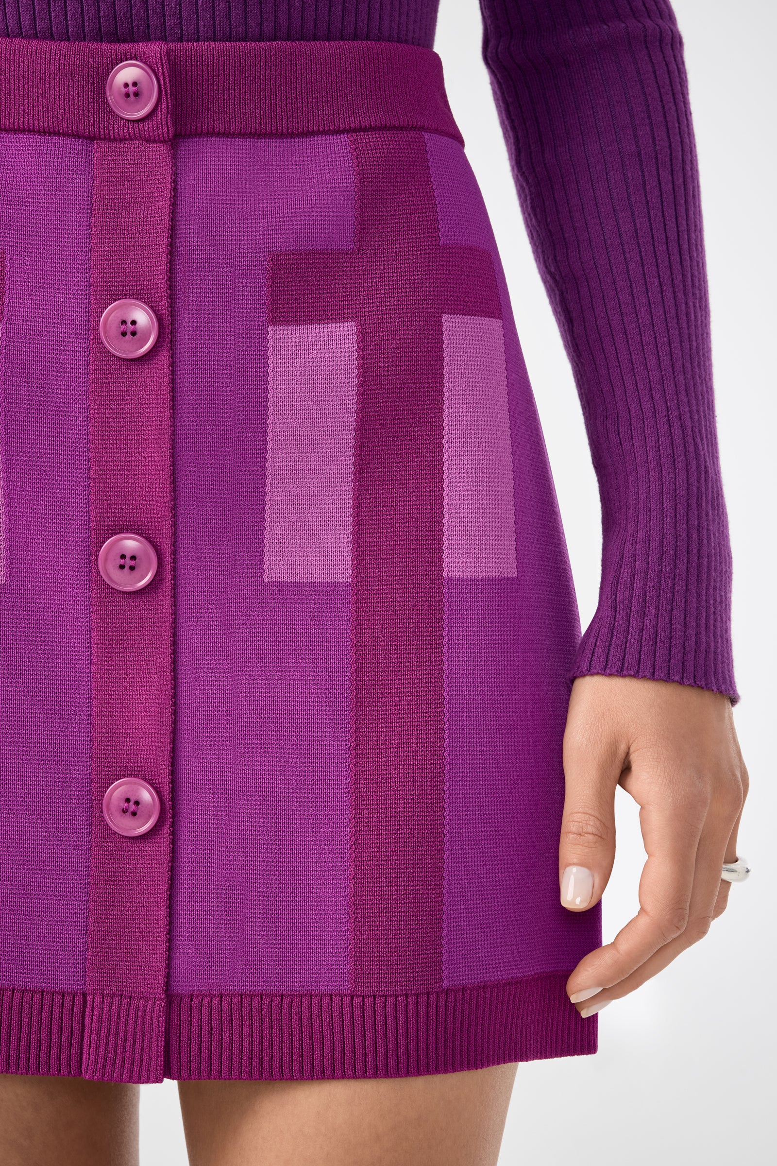 A person wearing the Artemis Skirt—a vibrant purple knit skirt with large front buttons and geometric patterns—paired with a ribbed long-sleeve top. Only the lower torso and one hand are visible.