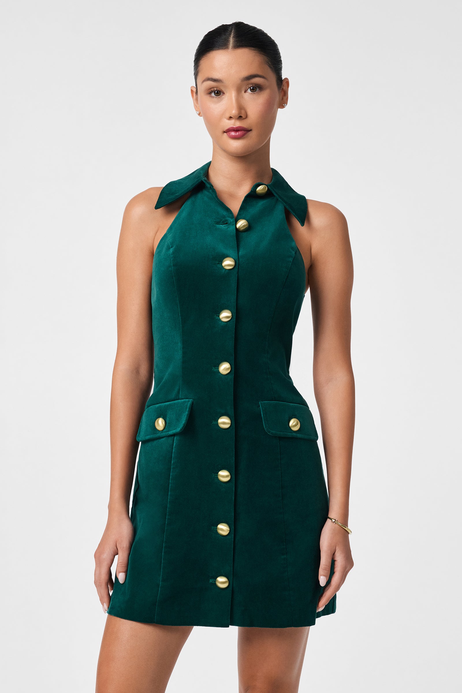 A woman models the Safina Dress, a sleeveless, collared dark green velvet dress with gold buttons and two front pockets, standing against a plain light background.