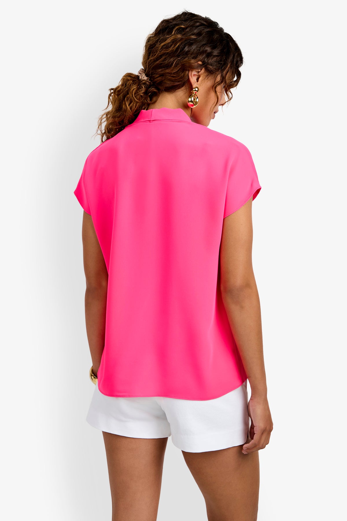 A woman with curly hair is seen from the back wearing the bright pink Turin Slash Tie Top and white shorts. She accessorizes with gold earrings and a bracelet, standing against a plain white background.
