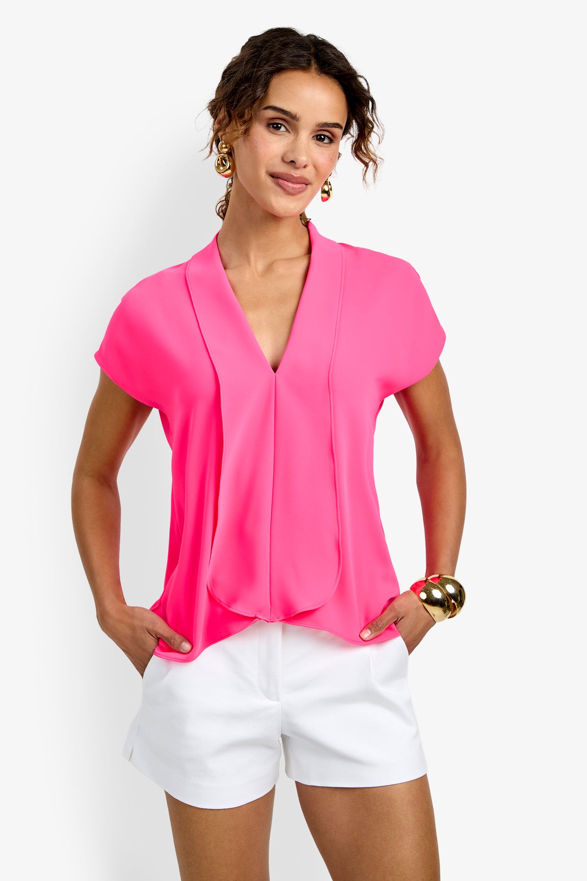 A woman stands with her hands in her pockets against a plain white background, wearing the bright pink Turin Slash Tie Top and white shorts, accessorized with gold hoop earrings and a chunky gold bracelet.