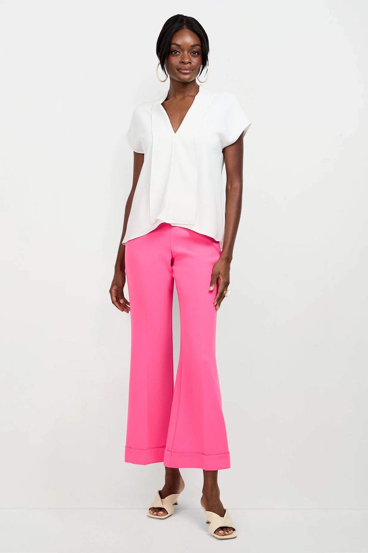 A woman stands against a white background wearing the Turin Slash Tie Top, bright pink wide-leg pants, white heeled sandals, and large hoop earrings. She smiles with her hands by her sides.