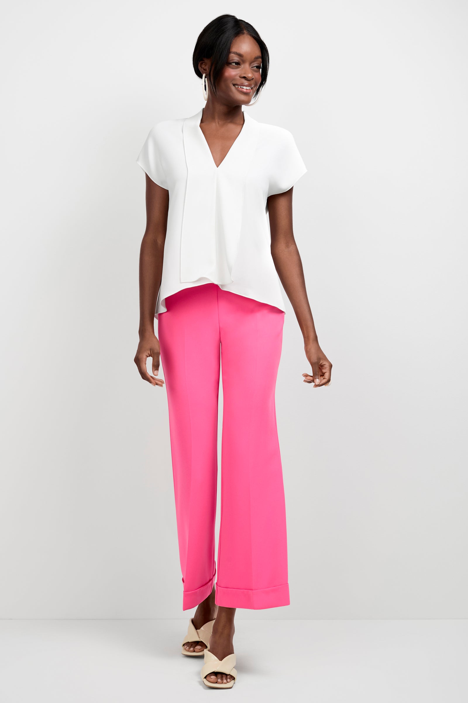A woman stands against a plain white background wearing the Turin Slash Tie Top, bright pink wide-leg trousers, and white heeled sandals. She is smiling and looking to the side.