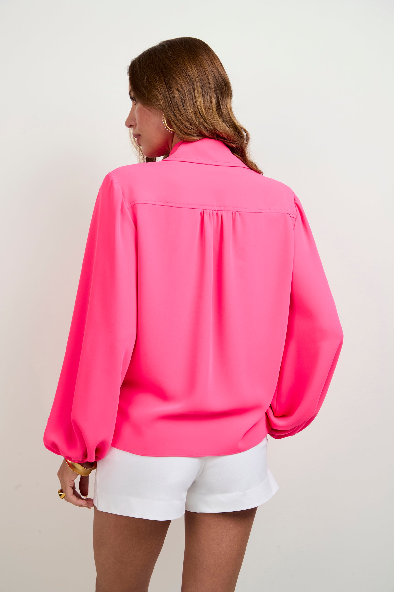 A person with long brown hair is seen from the back, wearing the Lambro Peasant Top in bright pink with puffy long sleeves and white shorts, set against a plain light background.