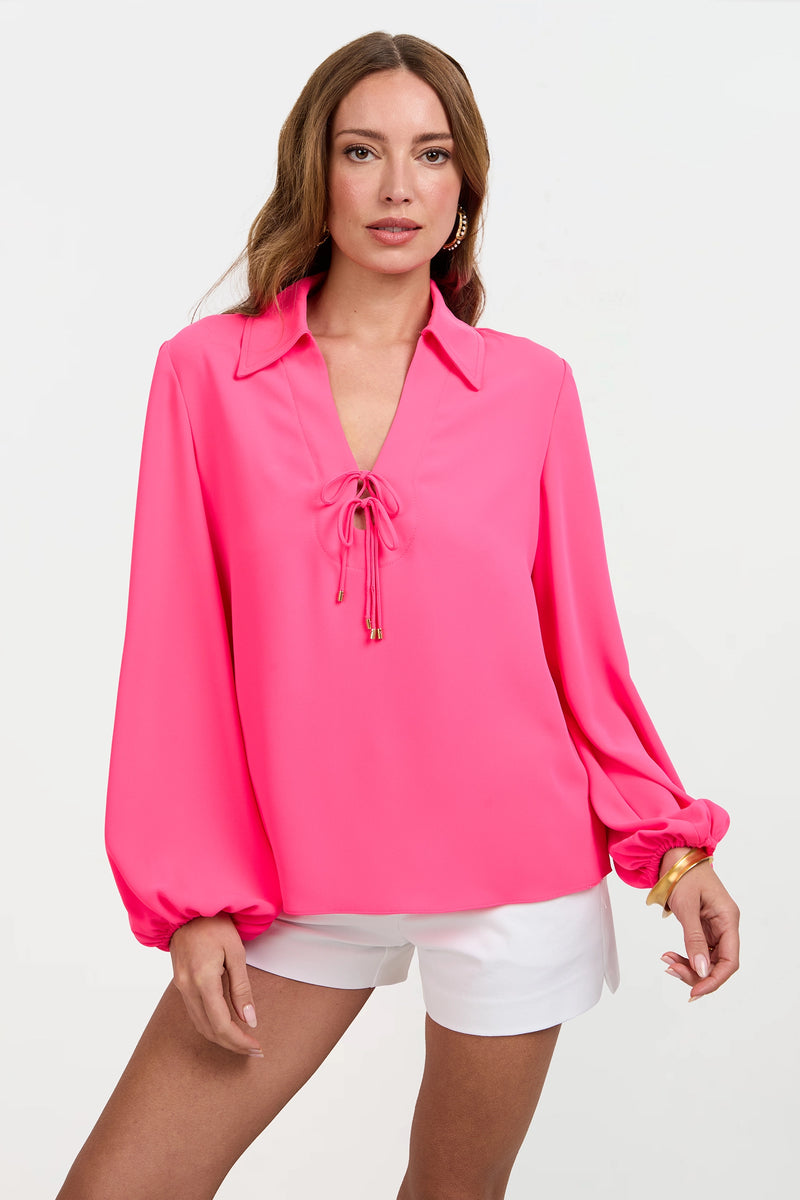 A woman models the Lambro Peasant Top, a bright pink long-sleeve blouse with a V-neck tie, paired with white shorts. She has long brown hair and accessorizes with gold earrings and a bracelet against a plain white background.