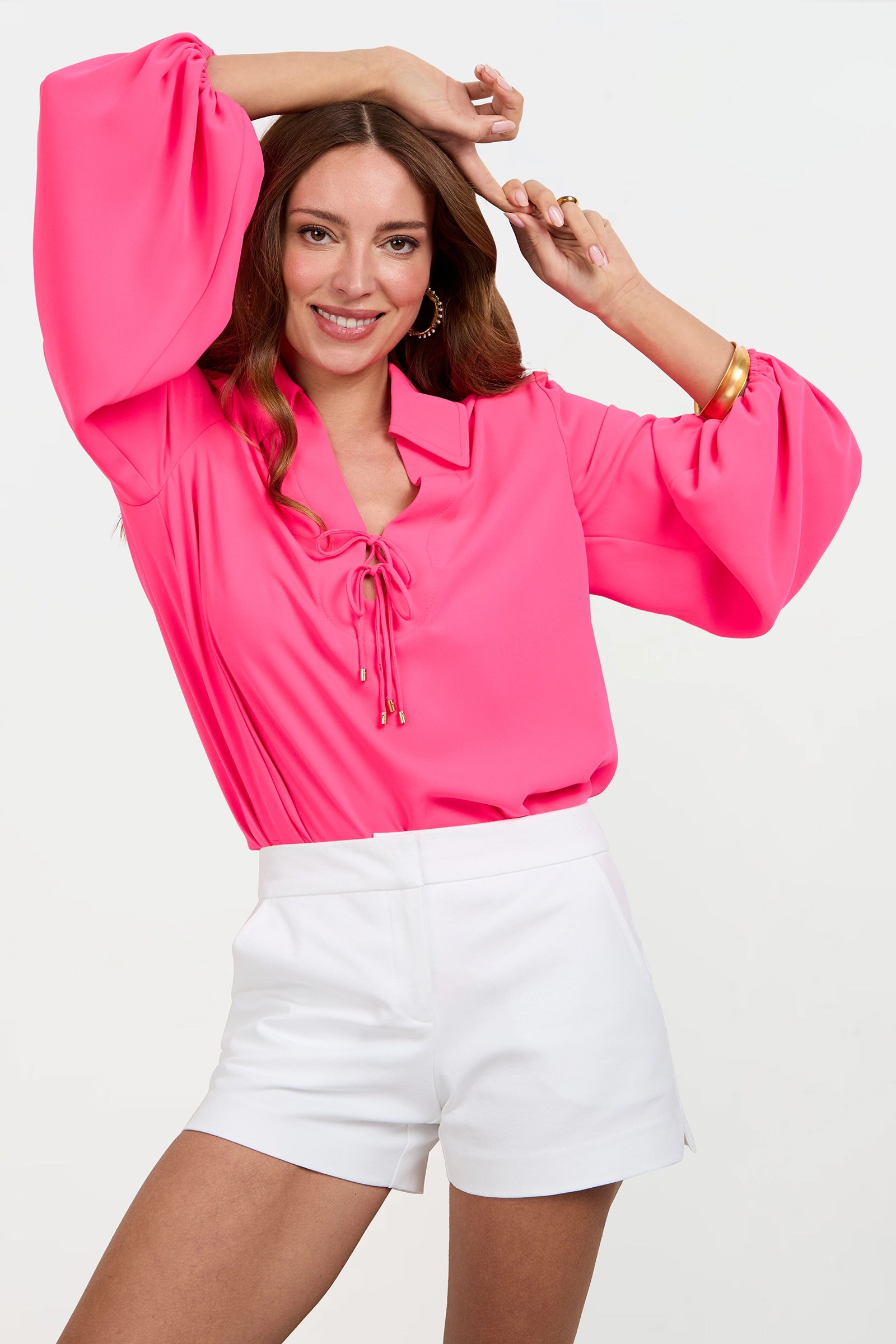 A woman with long brown hair and gold jewelry smiles while wearing the Lambro Peasant Top, a bright pink long-sleeve blouse with balloon sleeves, paired with white high-waisted shorts against a plain white background.