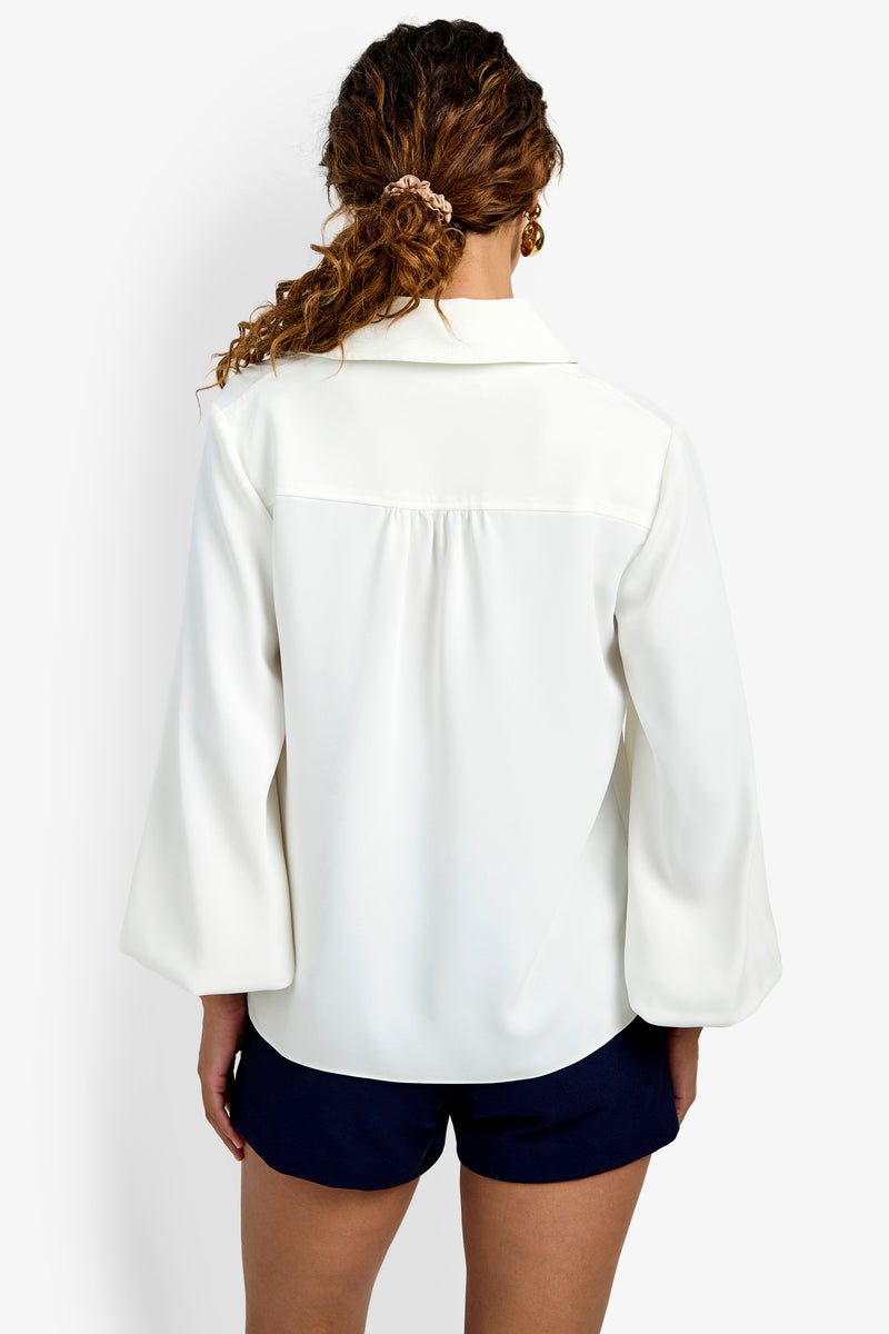 A woman with curly hair in a scrunchie is shown from the back, wearing the Lambro Peasant Top—a loose-fitting white blouse with long sleeves—paired with navy blue shorts against a plain white background.