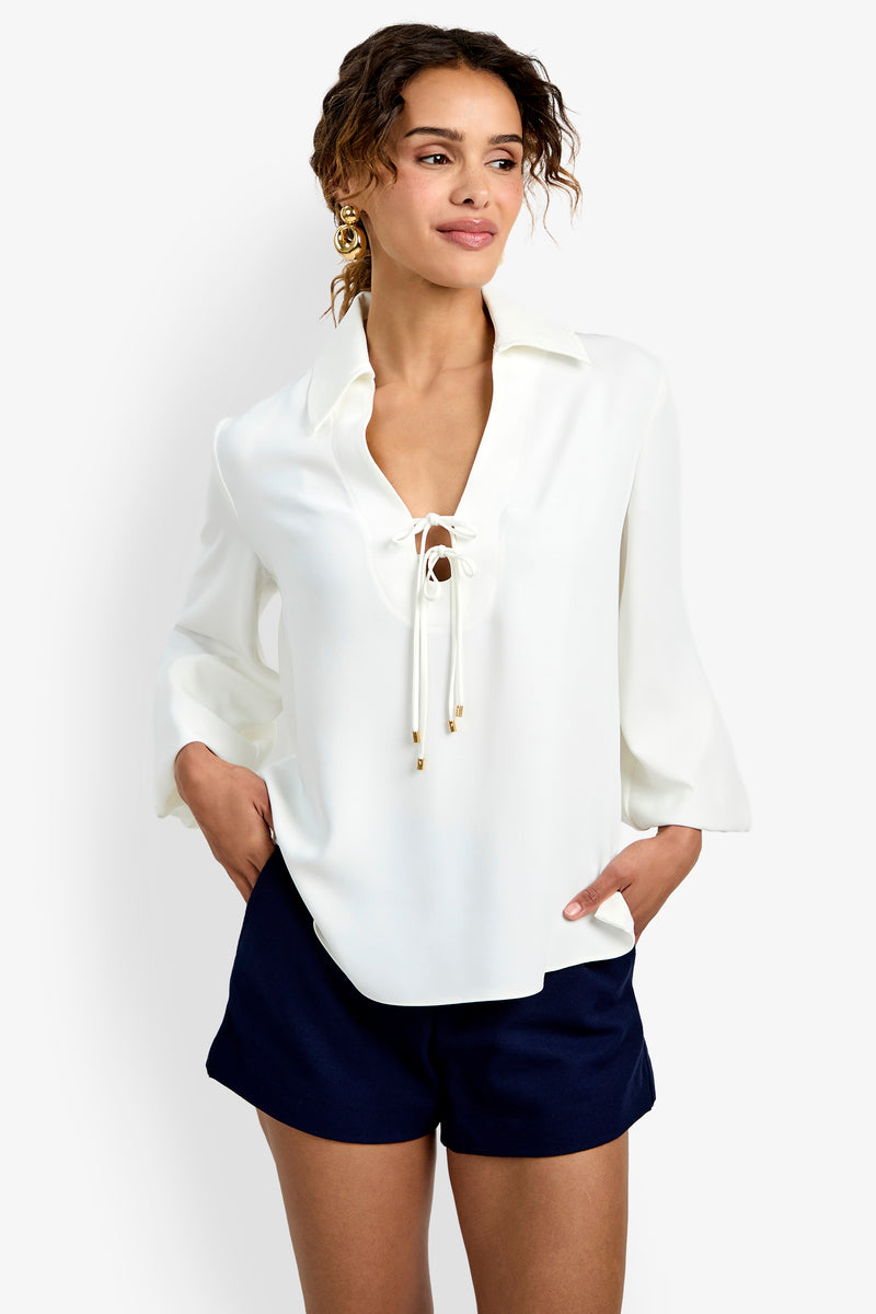 A woman wearing the Lambro Peasant Top—a white long-sleeve blouse with a lace-up neckline—paired with dark navy shorts stands with one hand in her pocket, looking off to the side and smiling.