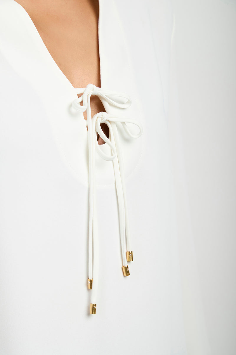 Close-up of the Lambro Peasant Top—a white blouse with a V-neckline, adorned with white tie strings featuring gold metal tips, tied in a bow at the chest.