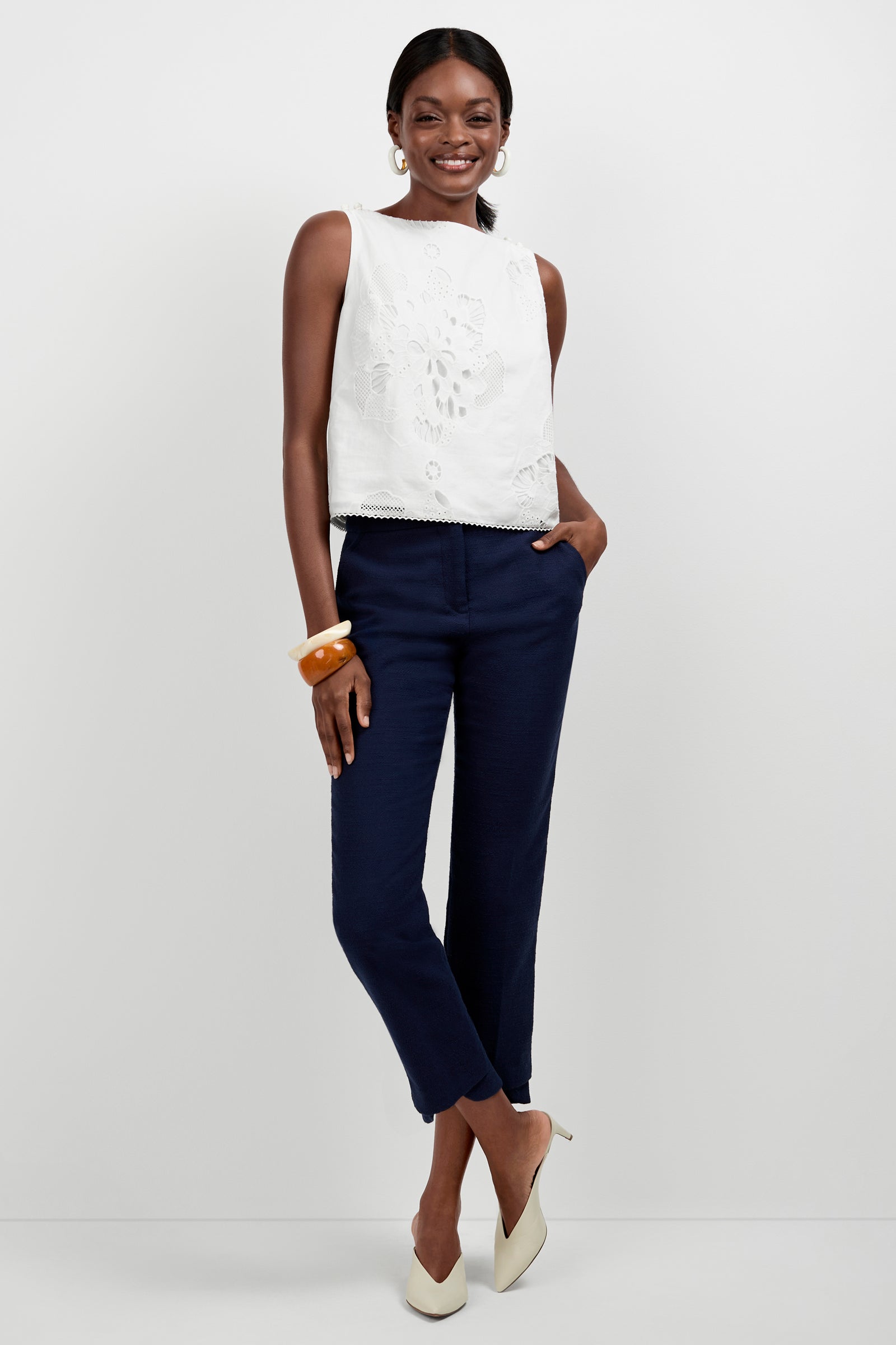 A woman smiles with her hands in her pockets, wearing dark navy Diffuse Pant, a sleeveless white embroidered top, cream heels, large hoop earrings, and chunky bracelets against a white background.