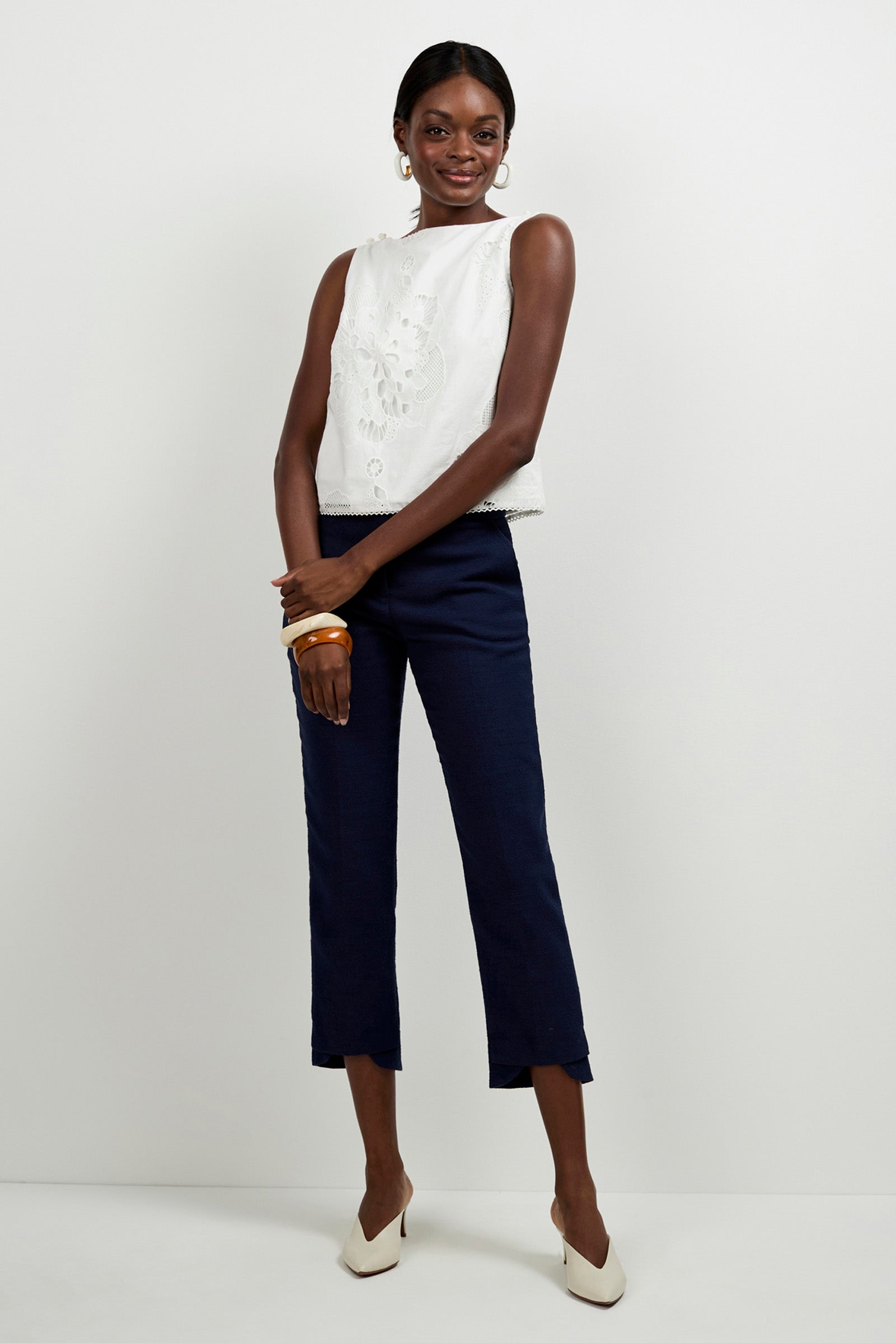 A woman stands against a plain background wearing Diffuse Pants, a sleeveless white embroidered top, white pointed heels, large hoop earrings, and wooden bangles, smiling gently with her arms crossed.
