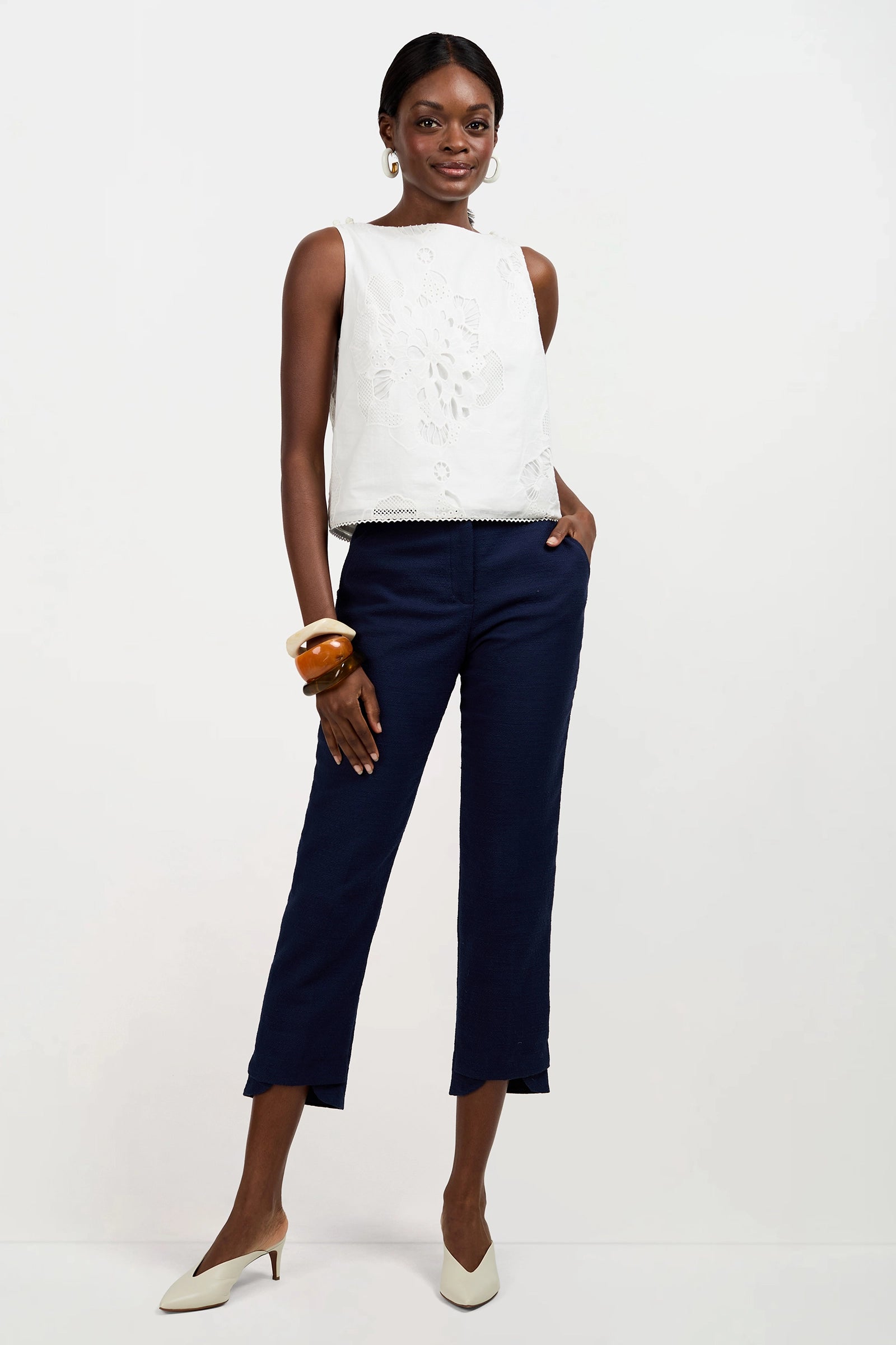 A woman smiles with her hands in her pockets, wearing the Alessandra Top in Whitewash, navy blue cropped pants, white heels, hoop earrings, and chunky bangles against a white background.