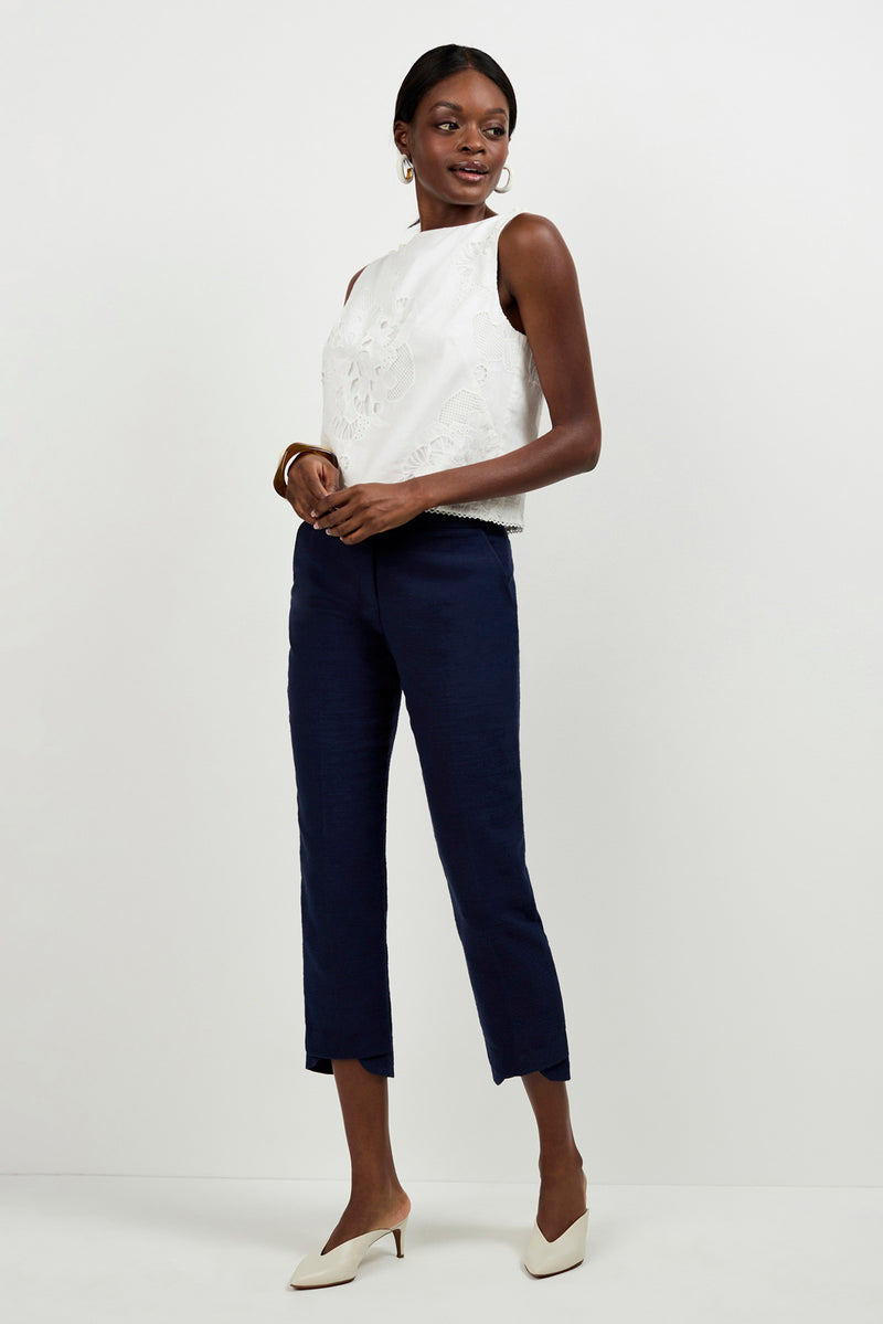 A woman smiles slightly and looks to the side against a white background, wearing Diffuse Pants in navy blue, a sleeveless white top with floral embroidery, white heeled shoes, hoop earrings, and a bracelet.