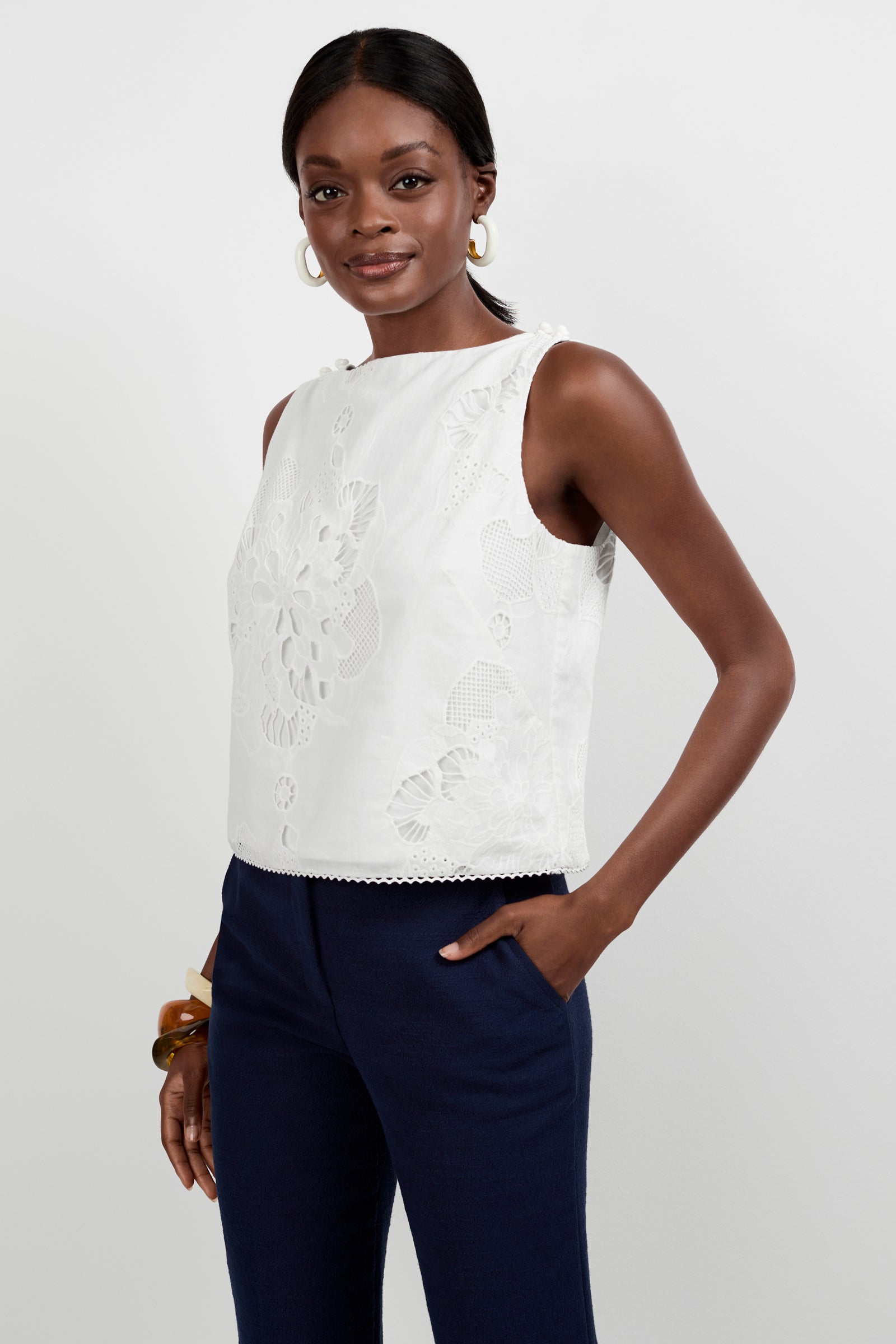 A woman models the Alessandra Top, a sleeveless white lace top with floral patterns, paired with navy blue pants and gold hoop earrings. She smiles softly with one hand in her pocket against a plain background.