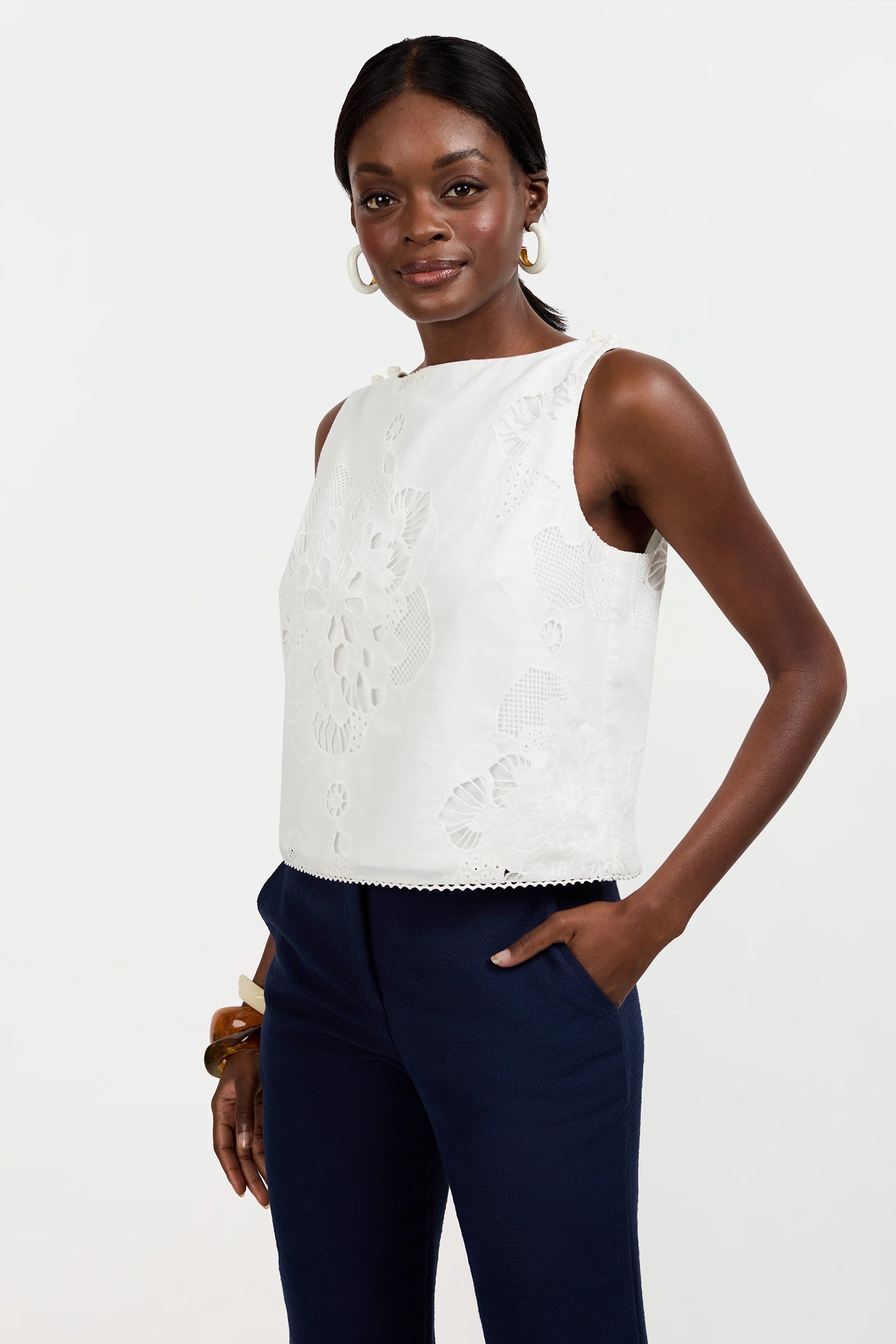 A woman models the Alessandra Top, a sleeveless white embroidered blouse, paired with navy blue pants. She stands with one hand in her pocket and smiles slightly against a plain white background.
