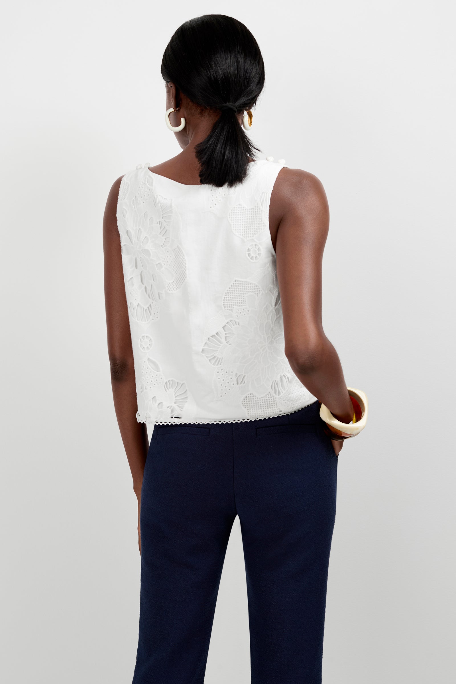 A woman with dark hair in a low ponytail, wearing the white sleeveless Alessandra Top and navy blue pants, faces away from the camera. She accessorizes with large hoop earrings and chunky bracelets on her left wrist.