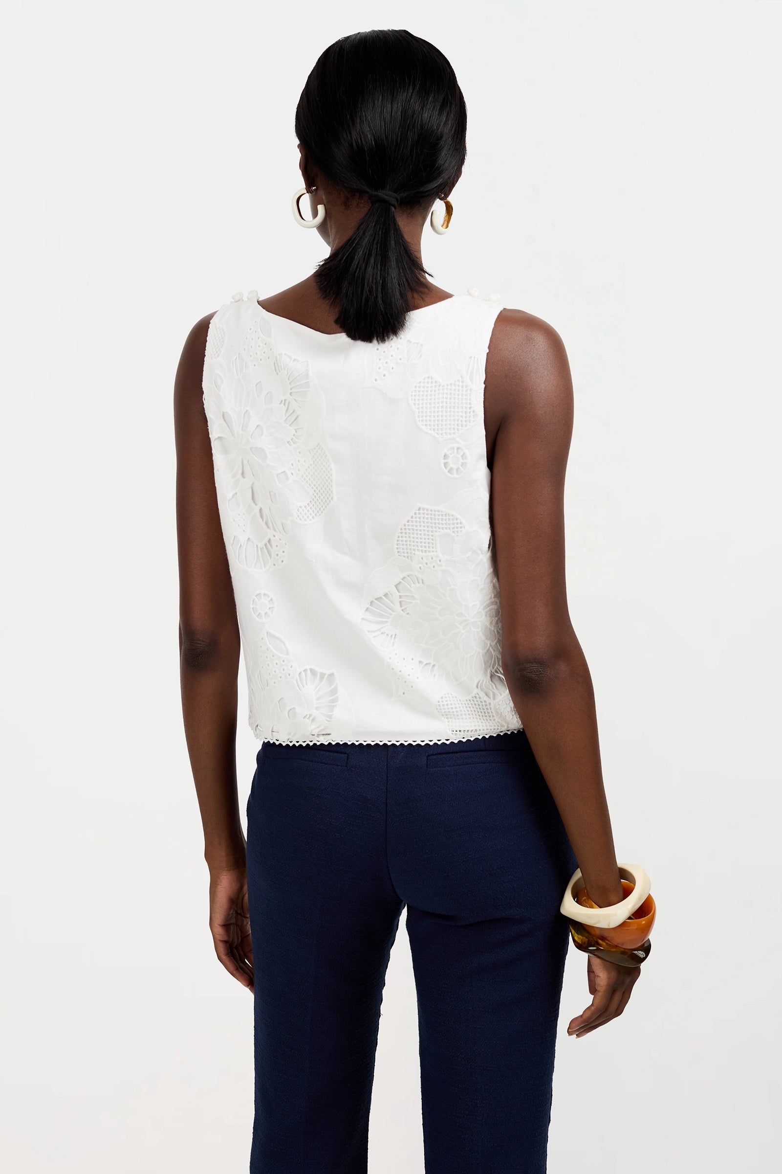 A woman is seen from behind wearing the sleeveless white lace Alessandra Top and navy blue pants, accessorized with hoop earrings and chunky bracelets on her left wrist. She has a low ponytail, and the background is plain white.