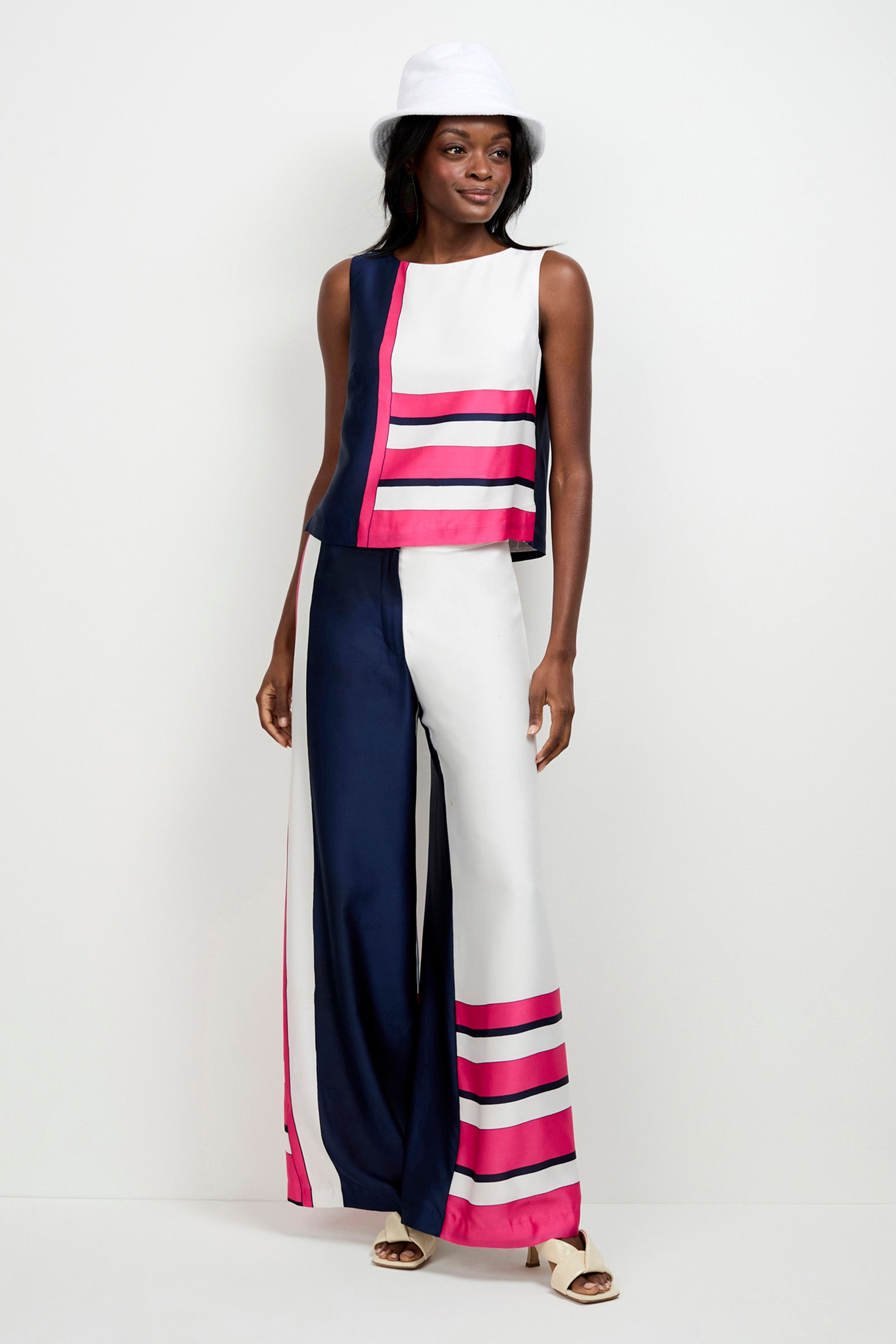 A woman wears the Cadorna Linee Sleeveless Top and wide-leg pants with navy, white, and pink stripes, paired with a white hat and sandals, standing against a plain background.