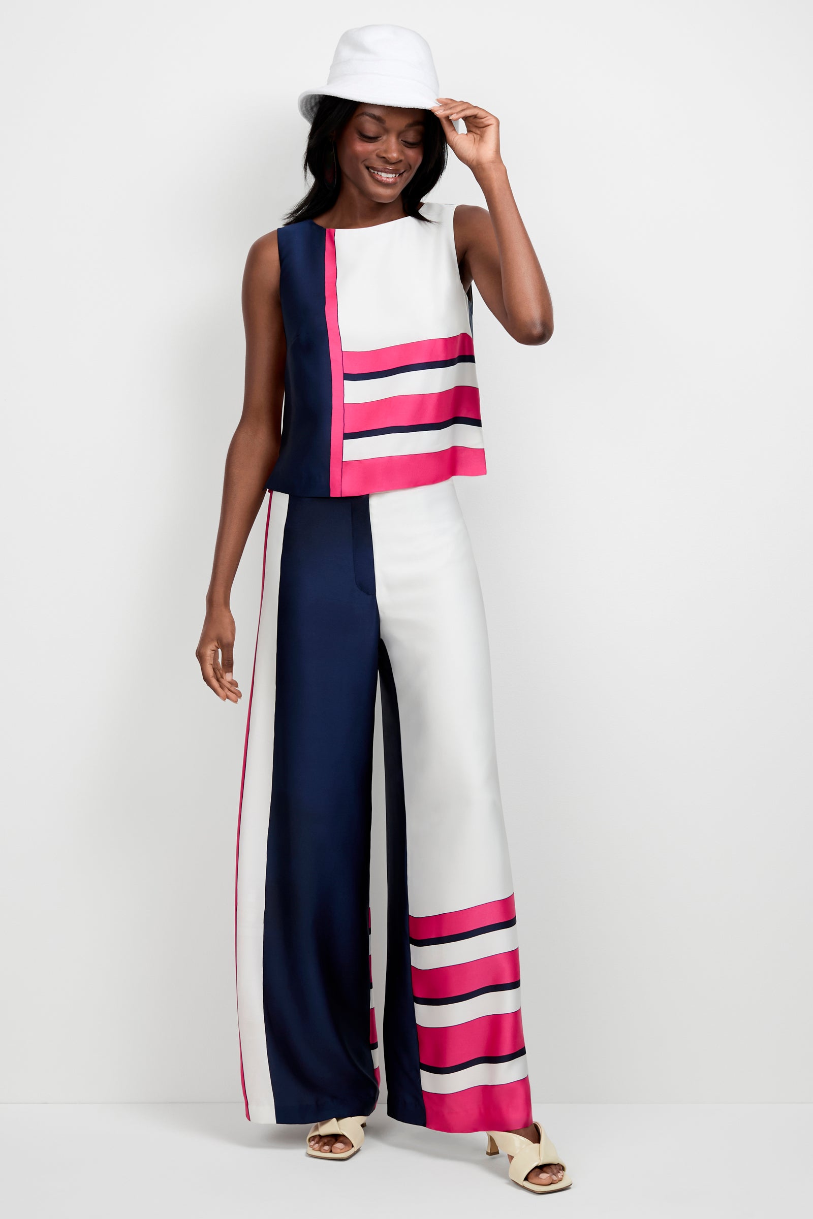 A woman smiles against a plain light background, wearing the Lanay Linee Wide-Leg Panta with bold navy, white, and pink geometric stripes, paired with a sleeveless top, white hat, and sandals.