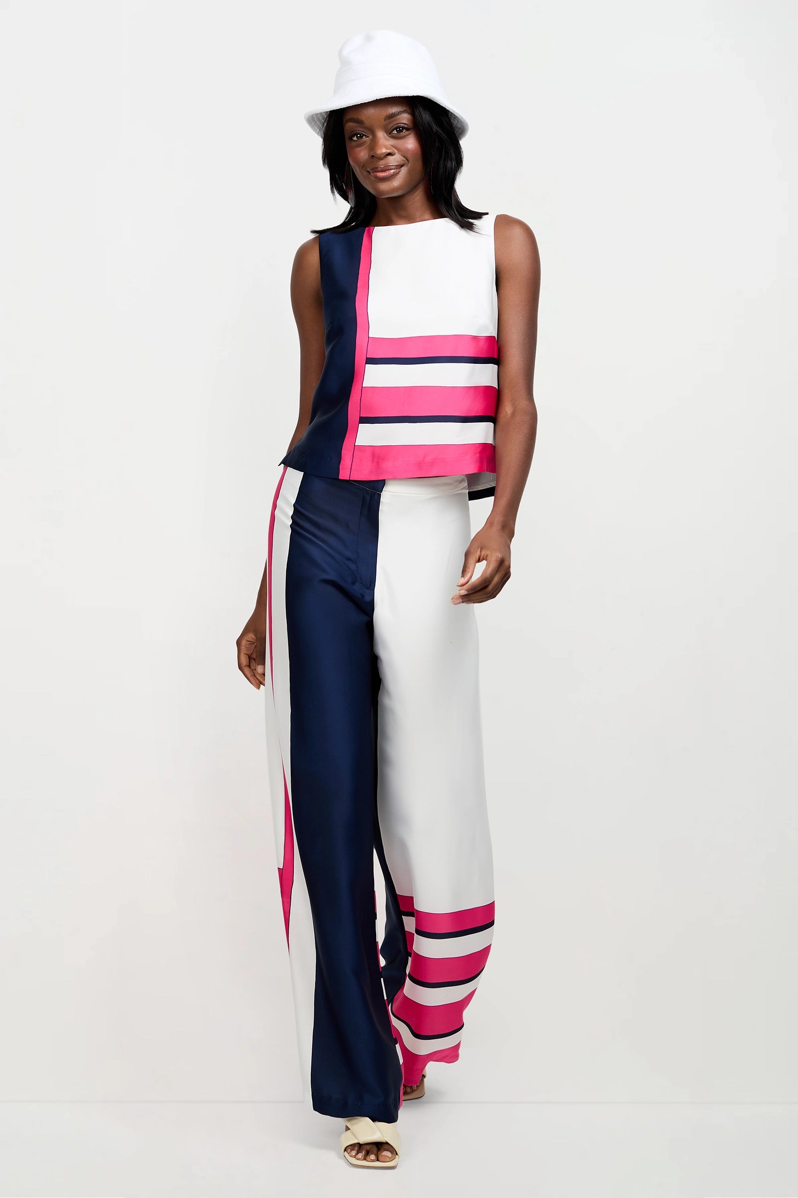 A woman stands smiling against a plain white background, wearing the Cadorna Linee Sleeveless Top and wide-leg pants featuring bold navy, white, and pink color-block stripes. She accessorizes with a white hat and open-toed shoes.