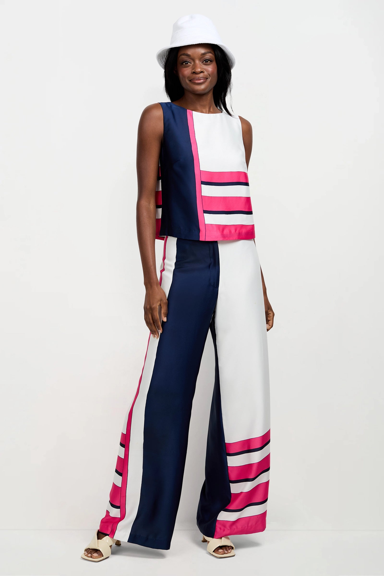 A woman wears the Cadorna Linee Sleeveless Top and wide-leg pants with bold navy, white, and pink stripes. She accessorizes with a white bucket hat and open-toe heels, standing against a plain white background.