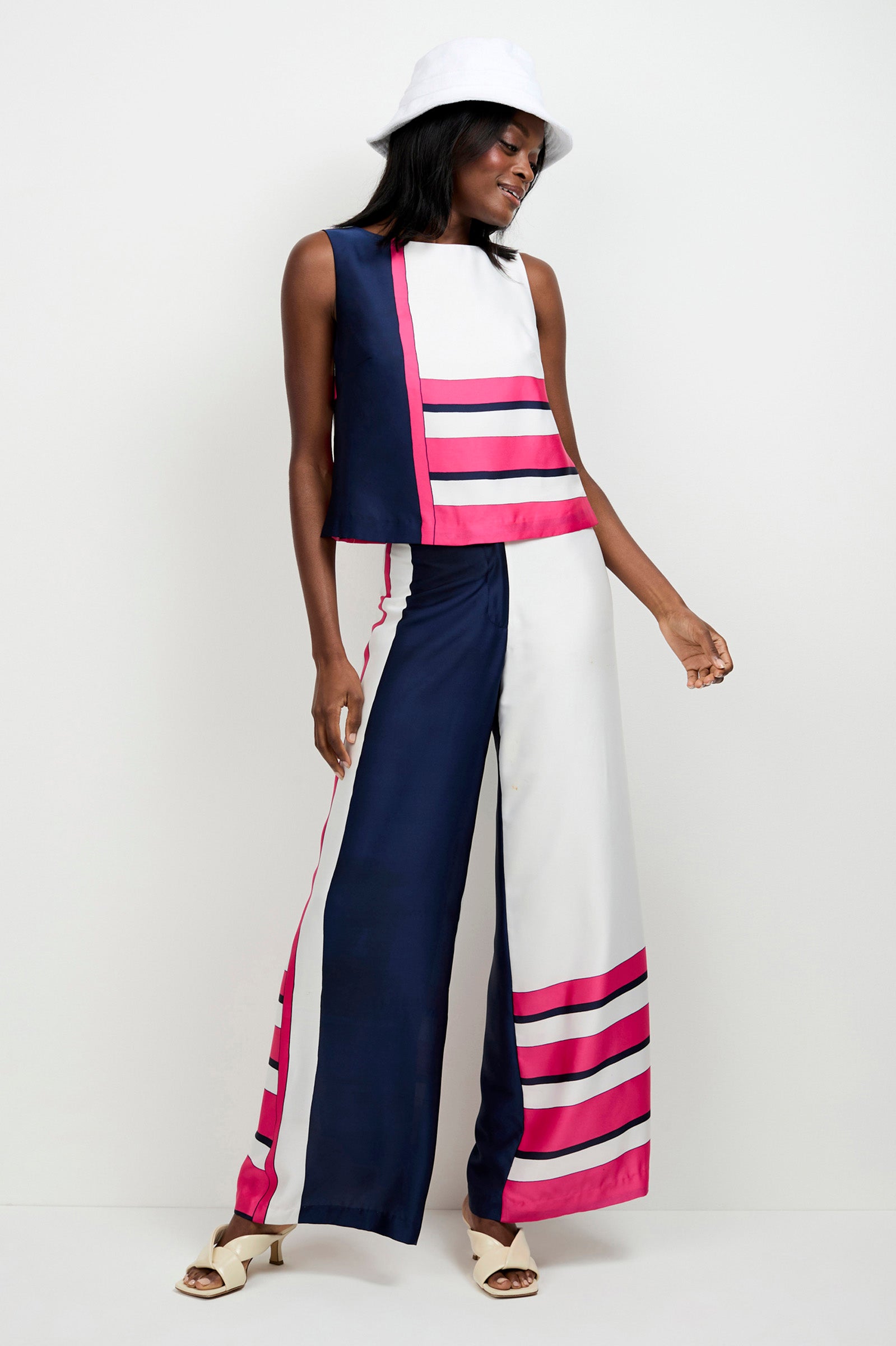 A woman poses against a plain white background wearing Lanay Linee Wide-Leg Pants with bold navy, white, and pink stripes, paired with a sleeveless top, a white bucket hat, and open-toe sandals.