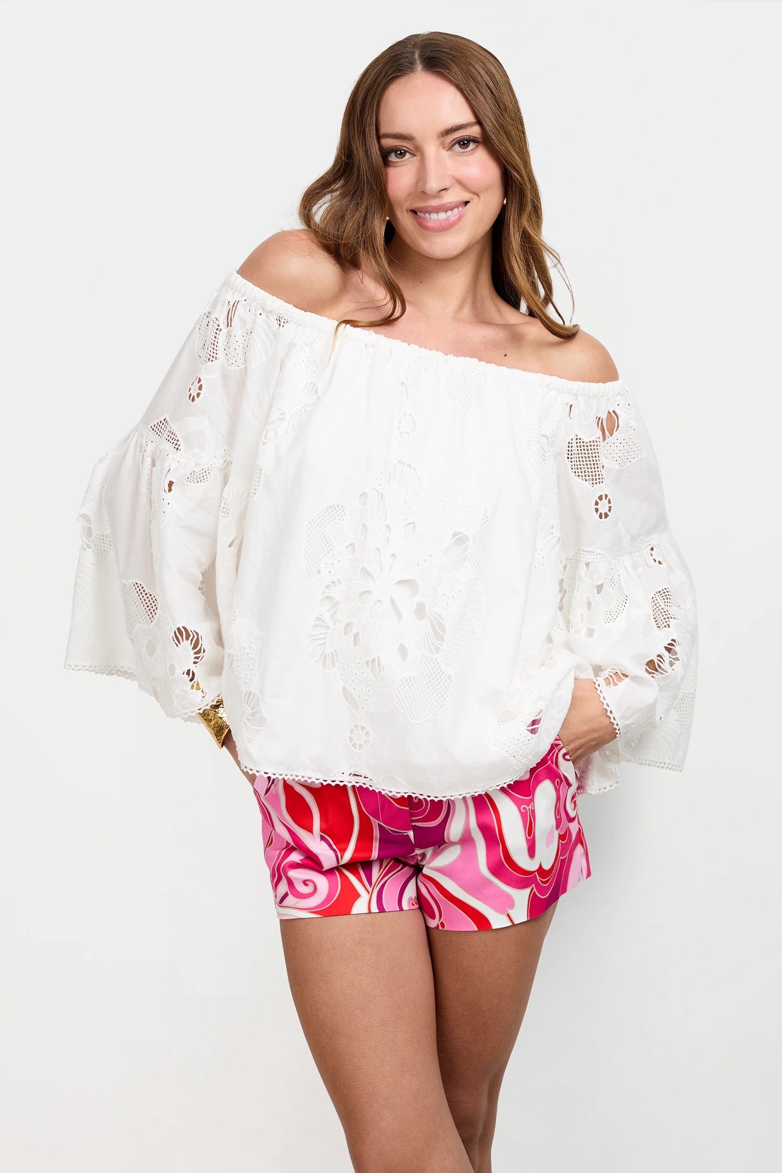 A woman smiles at the camera, wearing the Perry Off-Shoulder Top—an embroidered white blouse with bell sleeves—paired with colorful pink and red patterned shorts against a plain white background.