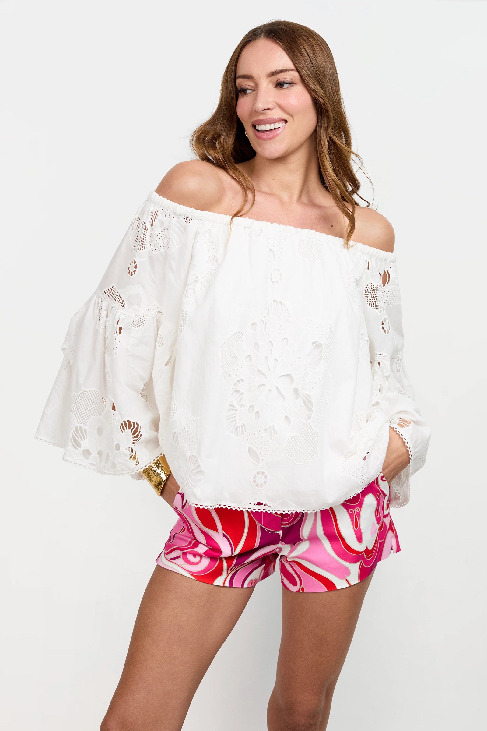 A woman smiles, wearing the Perry Off-Shoulder Top—white with eyelet floral cutouts and flared sleeves—paired with pink and red patterned shorts. She stands against a plain white background with one hand in her pocket.