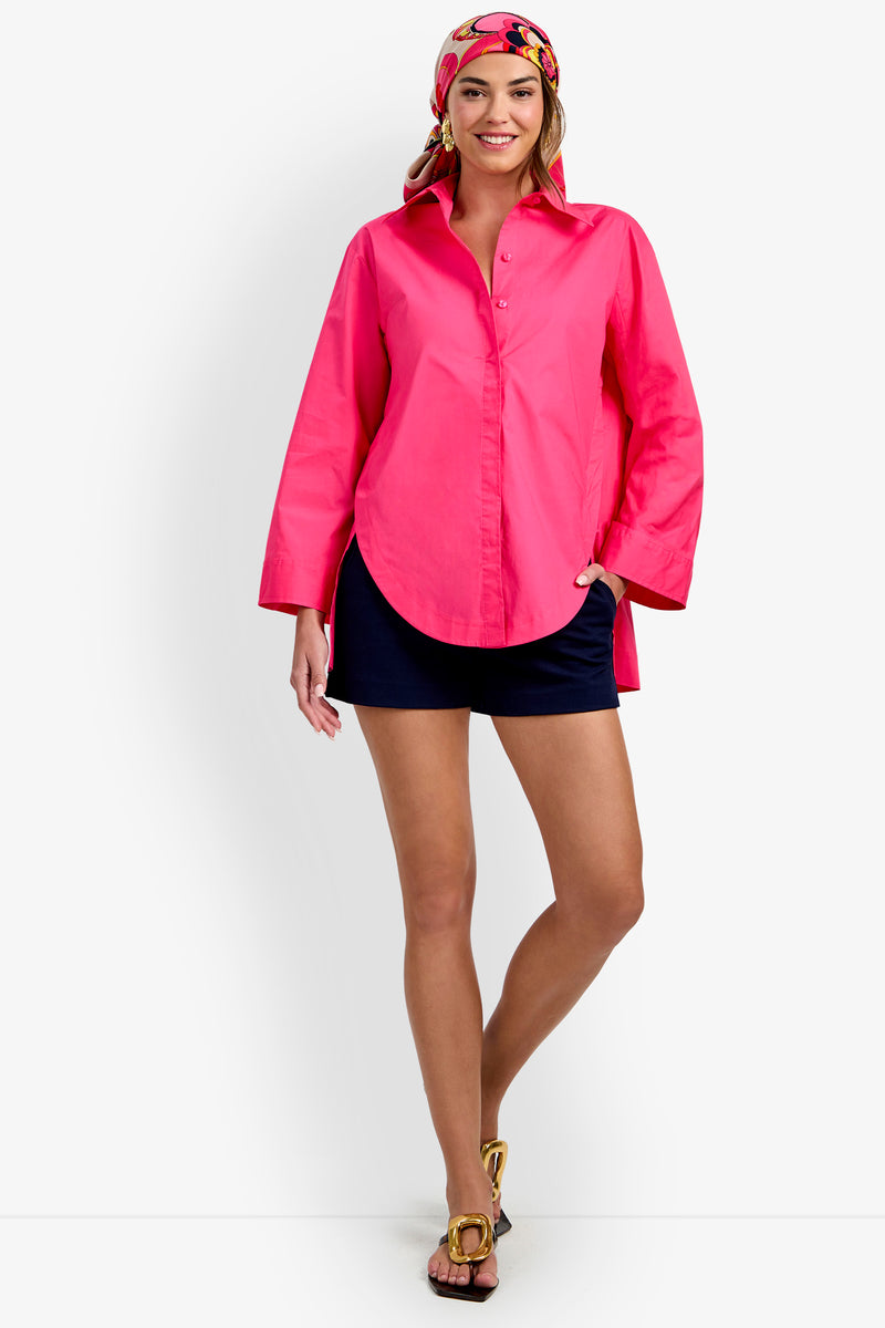 A woman smiles and stands against a white background, wearing the bright pink Olona Swing Top, navy shorts, a colorful headscarf, and gold-detailed sandals.