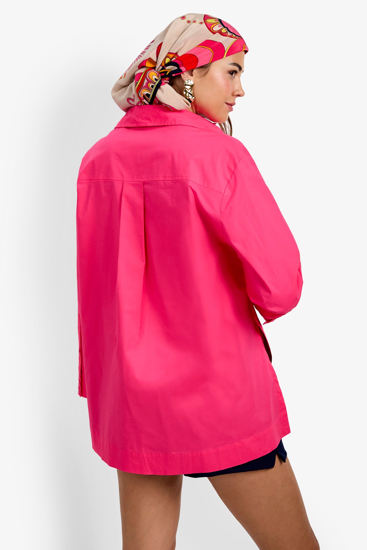 Wearing the Olona Swing Top in vibrant pink, paired with navy shorts, a patterned headscarf, and gold earrings, a woman stands facing away and looks over her shoulder against a plain white background.