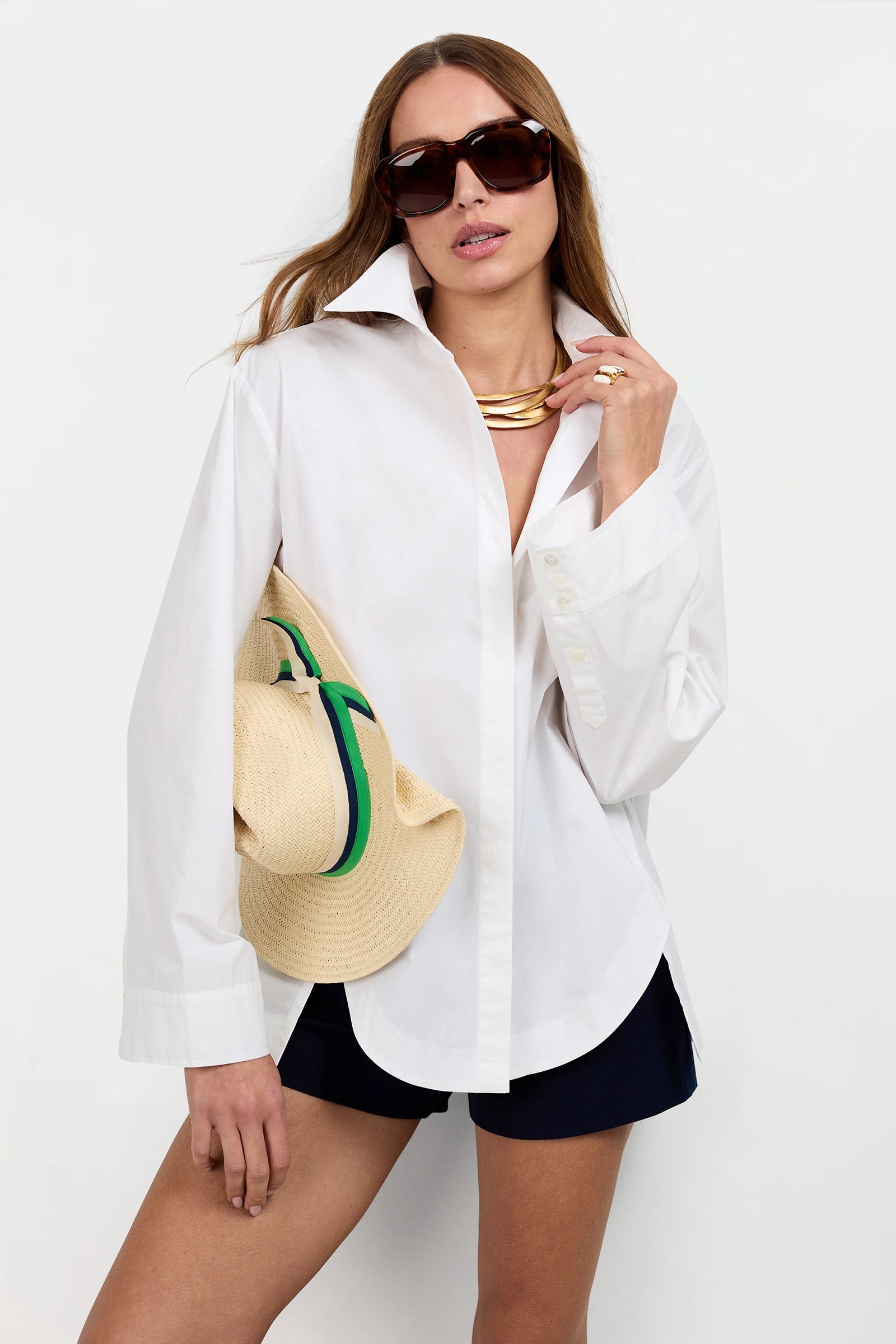 A woman models the Olona Swing Top—an oversized white button-up—paired with black shorts, sunglasses, a gold necklace, and a straw hat featuring green and blue bands, posing against a plain white background.