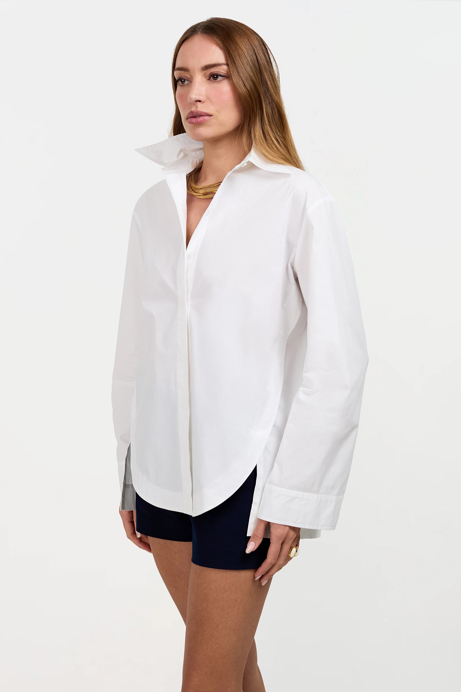 A woman with long brown hair wears the Olona Swing Top, a white button-up shirt with a high collar, paired with black shorts and gold jewelry, standing against a plain white background.