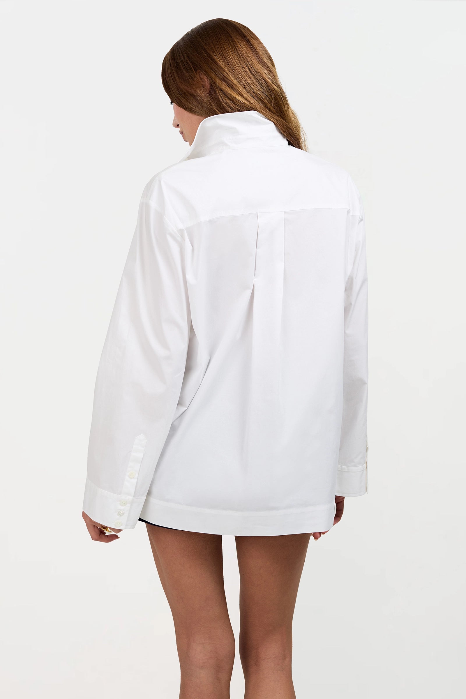 A person with long, light brown hair stands with their back to the camera, wearing the oversized Olona Swing Top—an airy white button-up shirt with long sleeves—set against a plain white background.