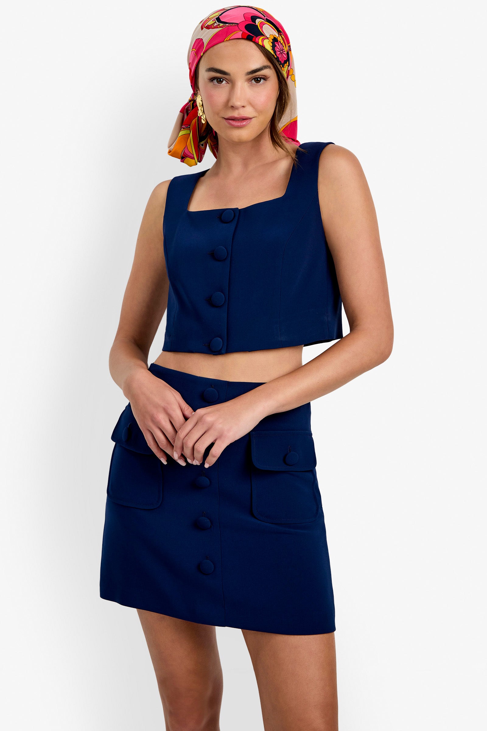 A woman wearing the Ramina Tailored Crop Vest in navy blue, styled with a matching skirt featuring front pockets and large buttons; she poses with her hands clasped and accessorizes with a vibrant pink and orange headscarf.