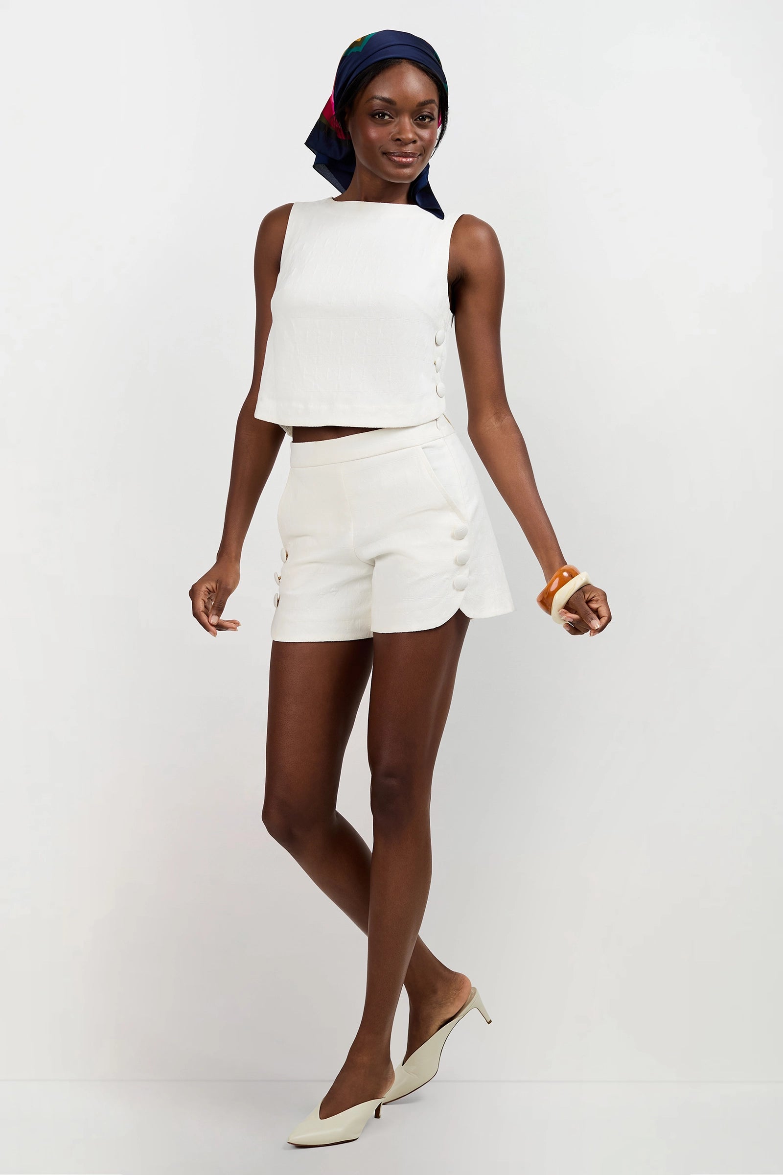 Wearing the Siara Button Side Top, high-waisted matching shorts with side buttons, a navy headscarf, white pointed heels, and a chunky bracelet, a woman poses and smiles against a plain white background.
