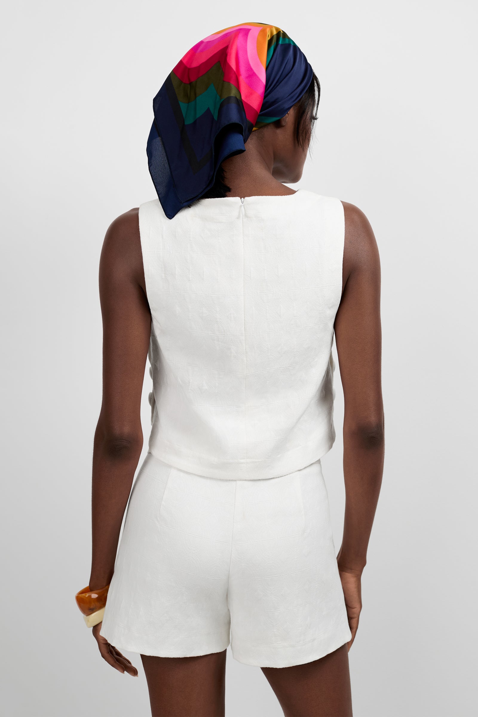 A person stands facing away, wearing the Siara Button Side Top in white with matching shorts. They have a colorful geometric headscarf and a chunky bracelet. The background is plain and light.