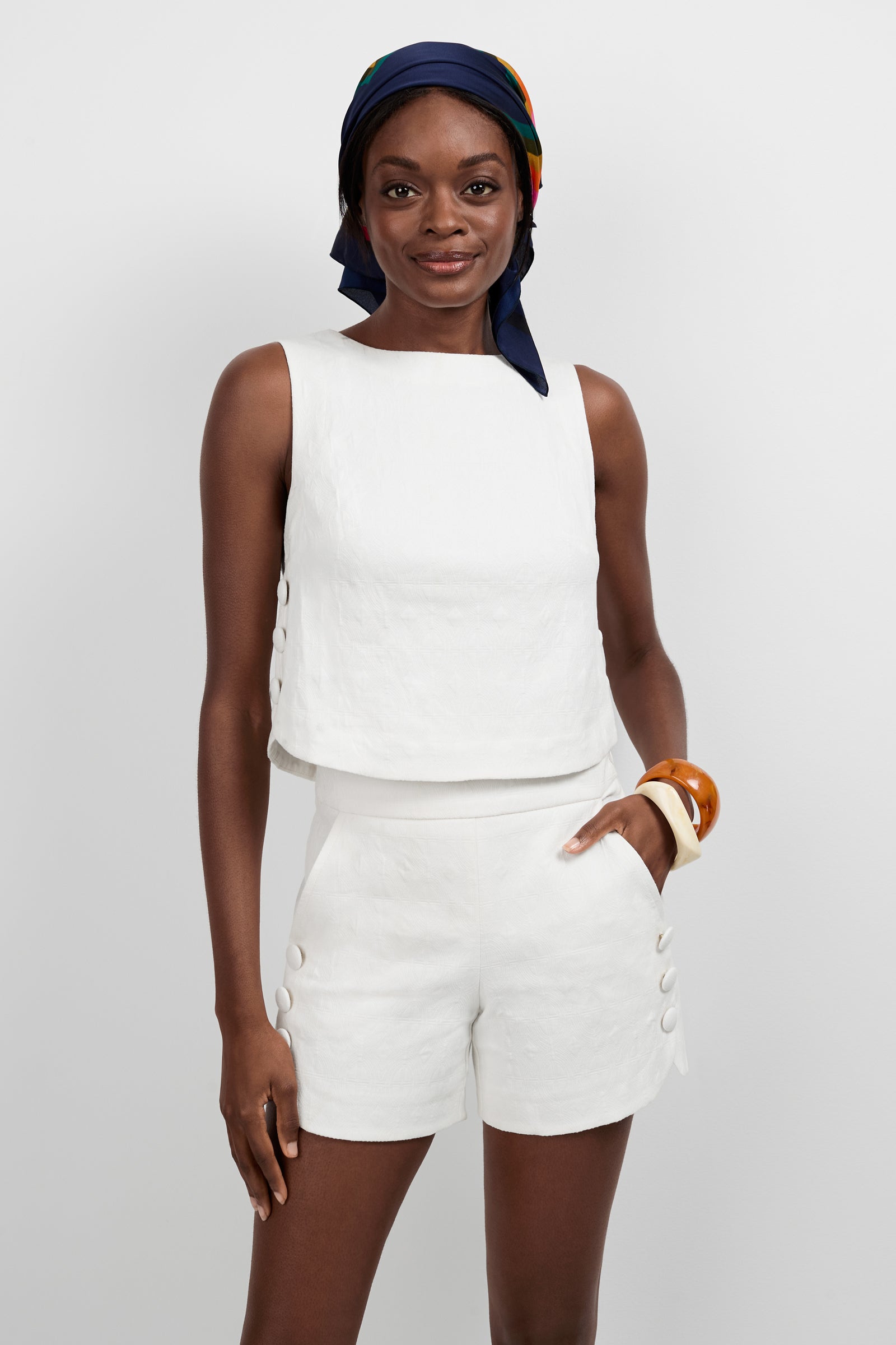 A woman wears the Siara Button Side Top paired with matching white shorts, standing against a plain background. She accessorizes with a colorful scarf and chunky bracelets, smiling softly at the camera.
