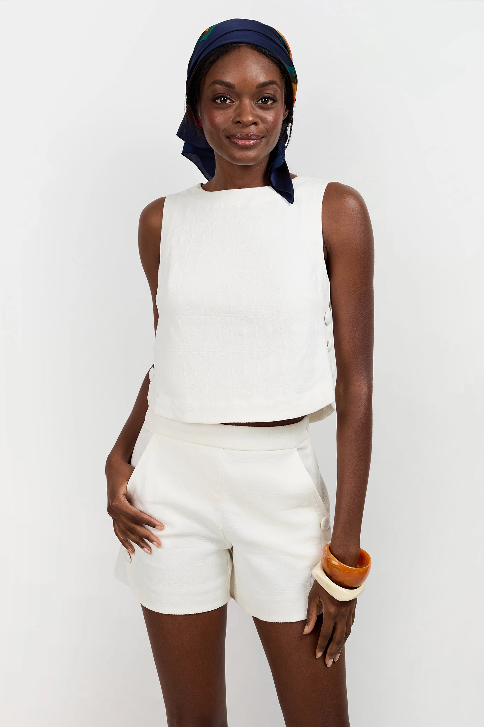 A woman models the Siara Button Side Top with matching shorts, one hand in her pocket. She accessorizes with a colorful headscarf and chunky bangles, posing against a plain white background.